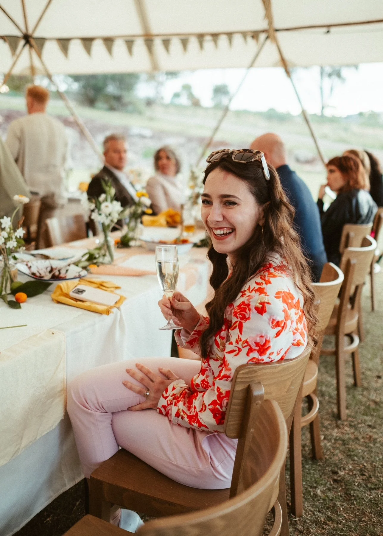 A woman with long brown hair, wearing a floral blouse and pink pants, sitting at a decorated outdoor event table under a tent, holding a glass of champagne and smiling.