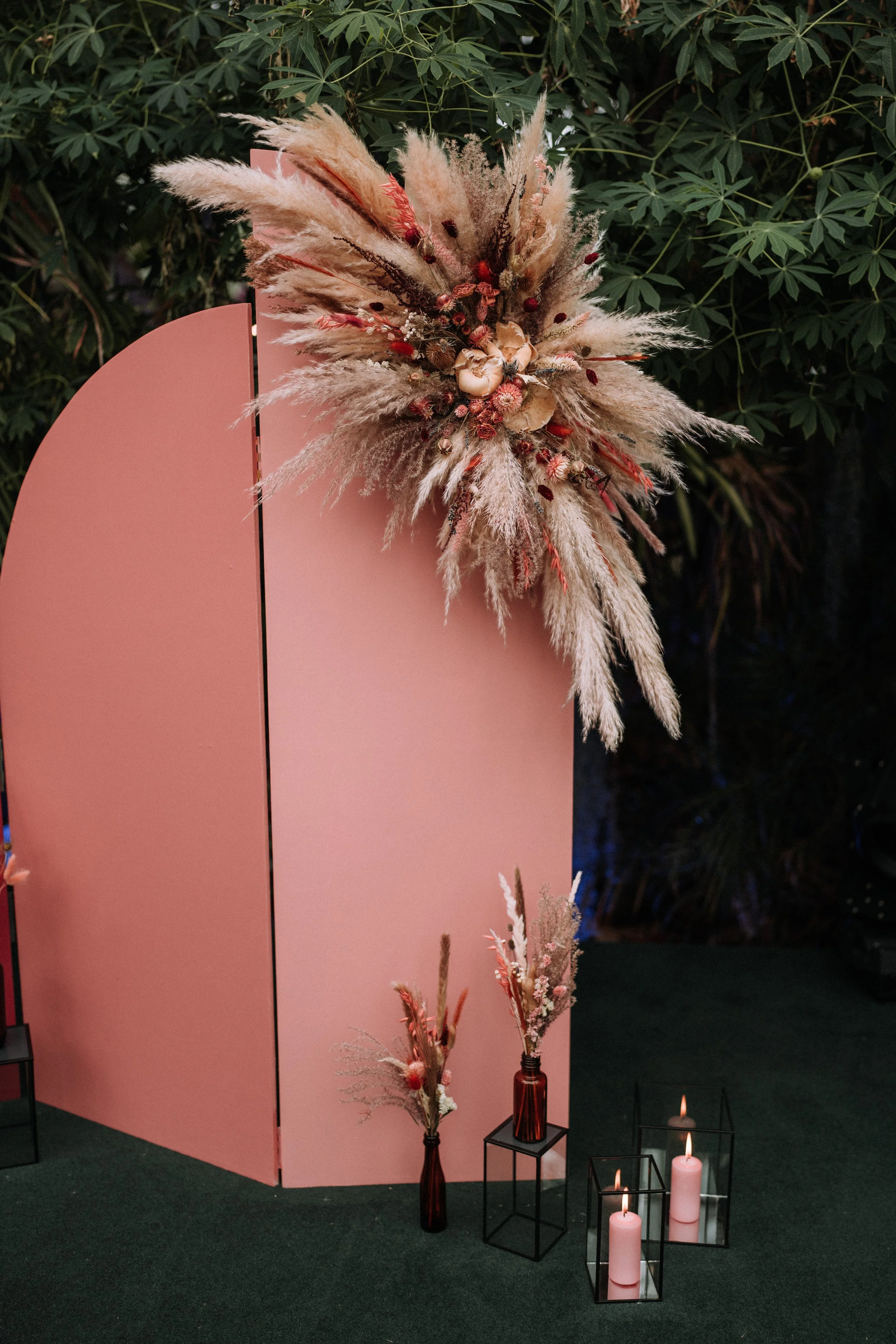 Decorative display featuring a pink partition, a vase with dried flowers including pampas grass and other dried floral elements, and lit pink candles in black metal candle holders.