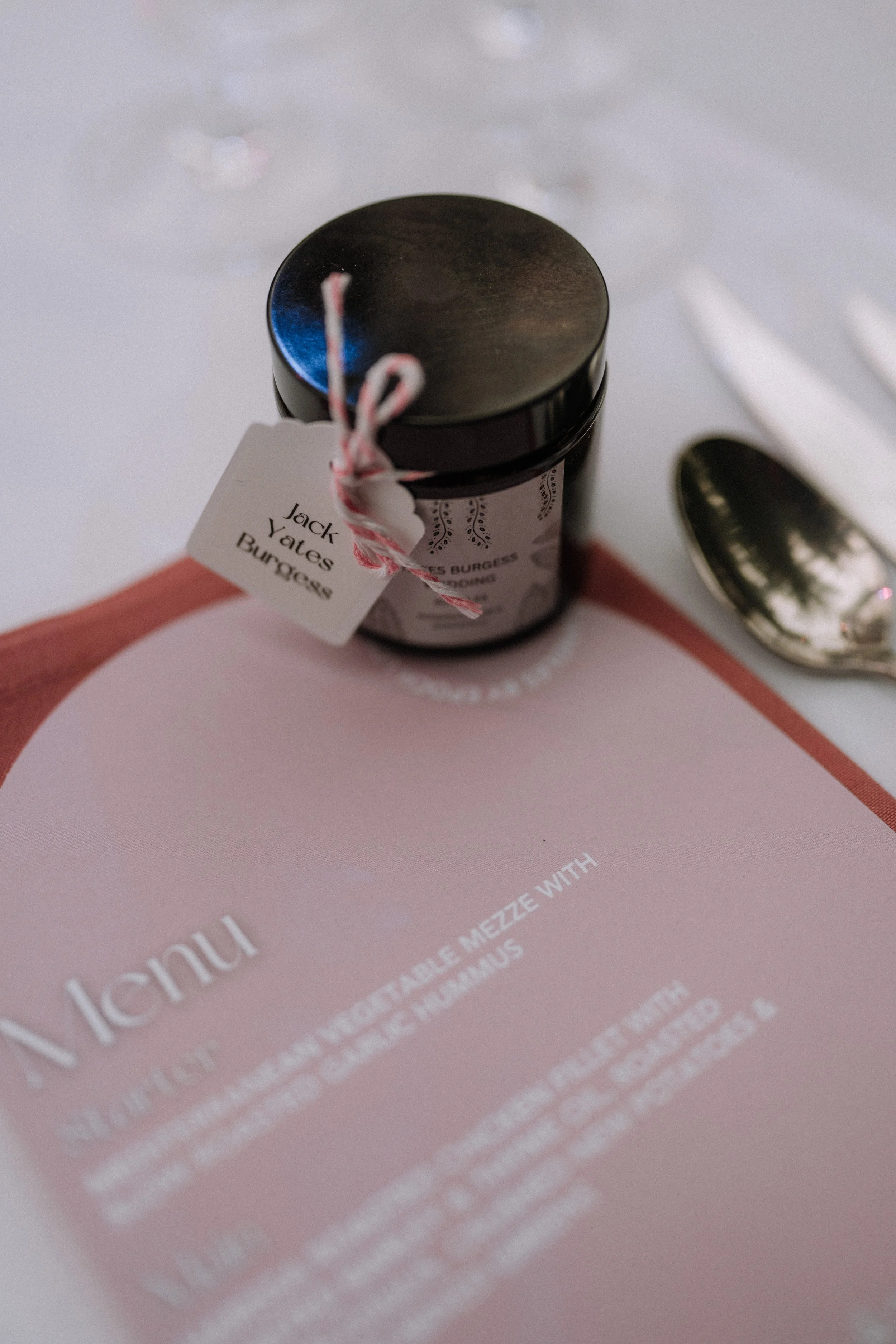A small jar with a black lid and a white label tied with red and white string. The label reads 'Jack Yates Burgess.' The jar is placed on a pink background, with a reflective metallic object partially visible on the right side.