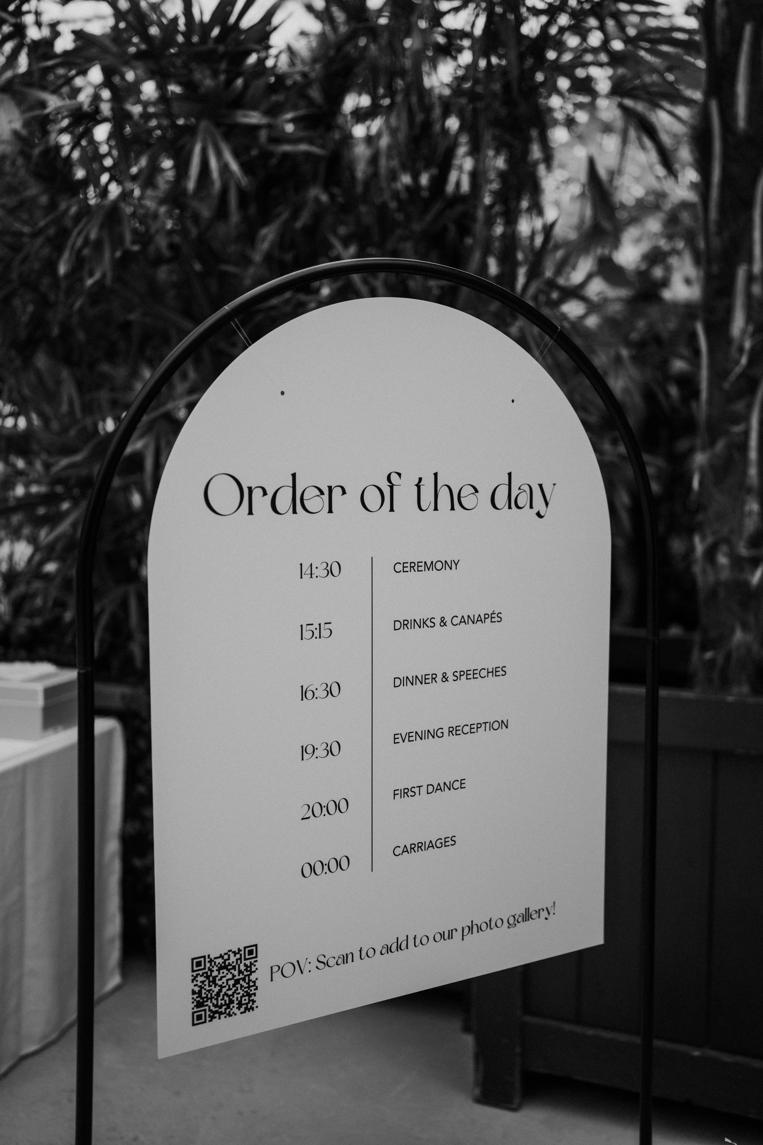 A wedding schedule sign displayed on a stand with a list of events including ceremony, drinks and canapés, dinner and speeches, evening reception, first dance, and carriages, with times from 14:30 to midnight on a white background with black text.