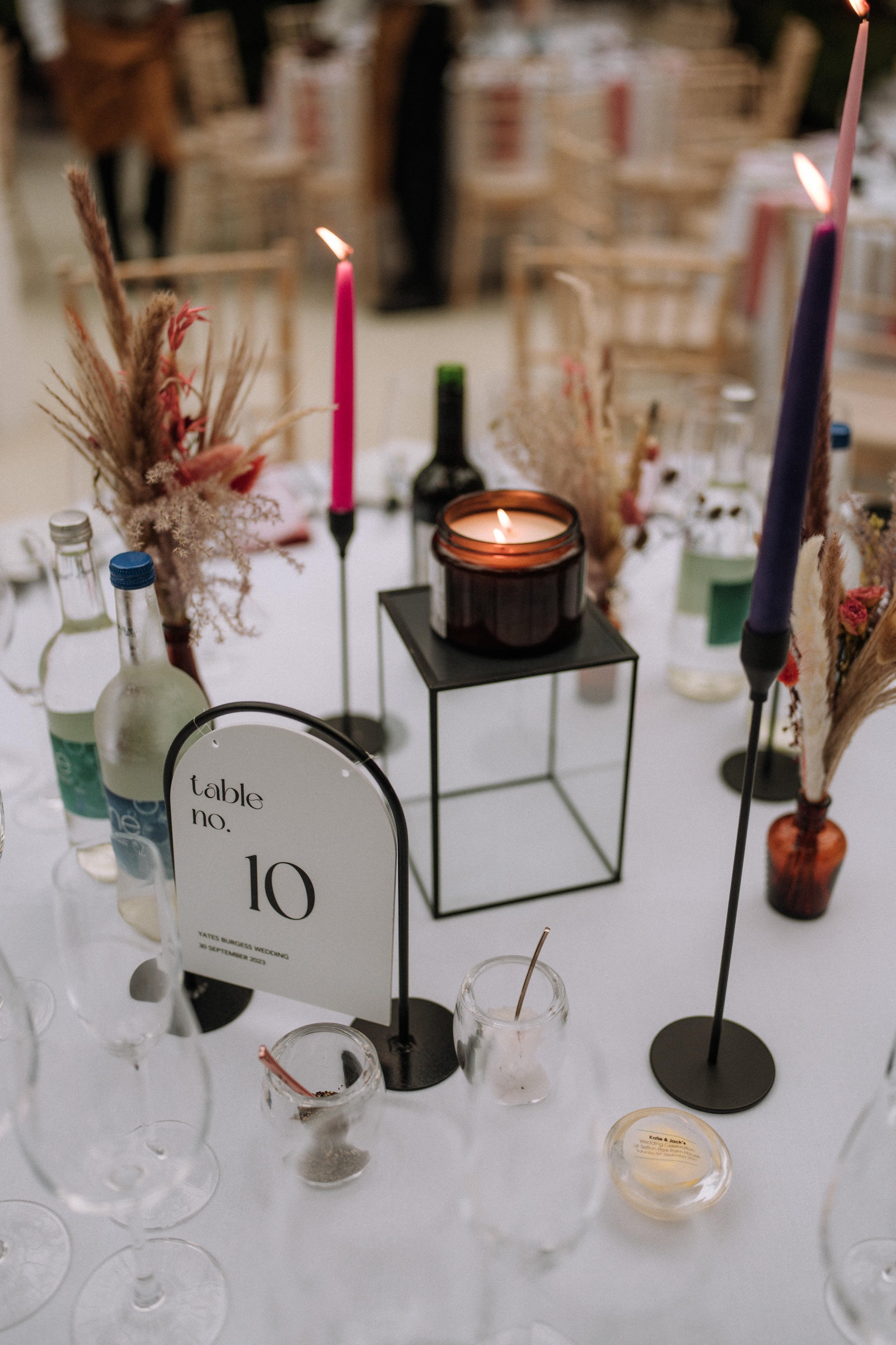 A wedding reception table setup with candles, dried flowers, empty bottles, and a table number sign indicating table no. 10, with blurred chairs and guests in the background.