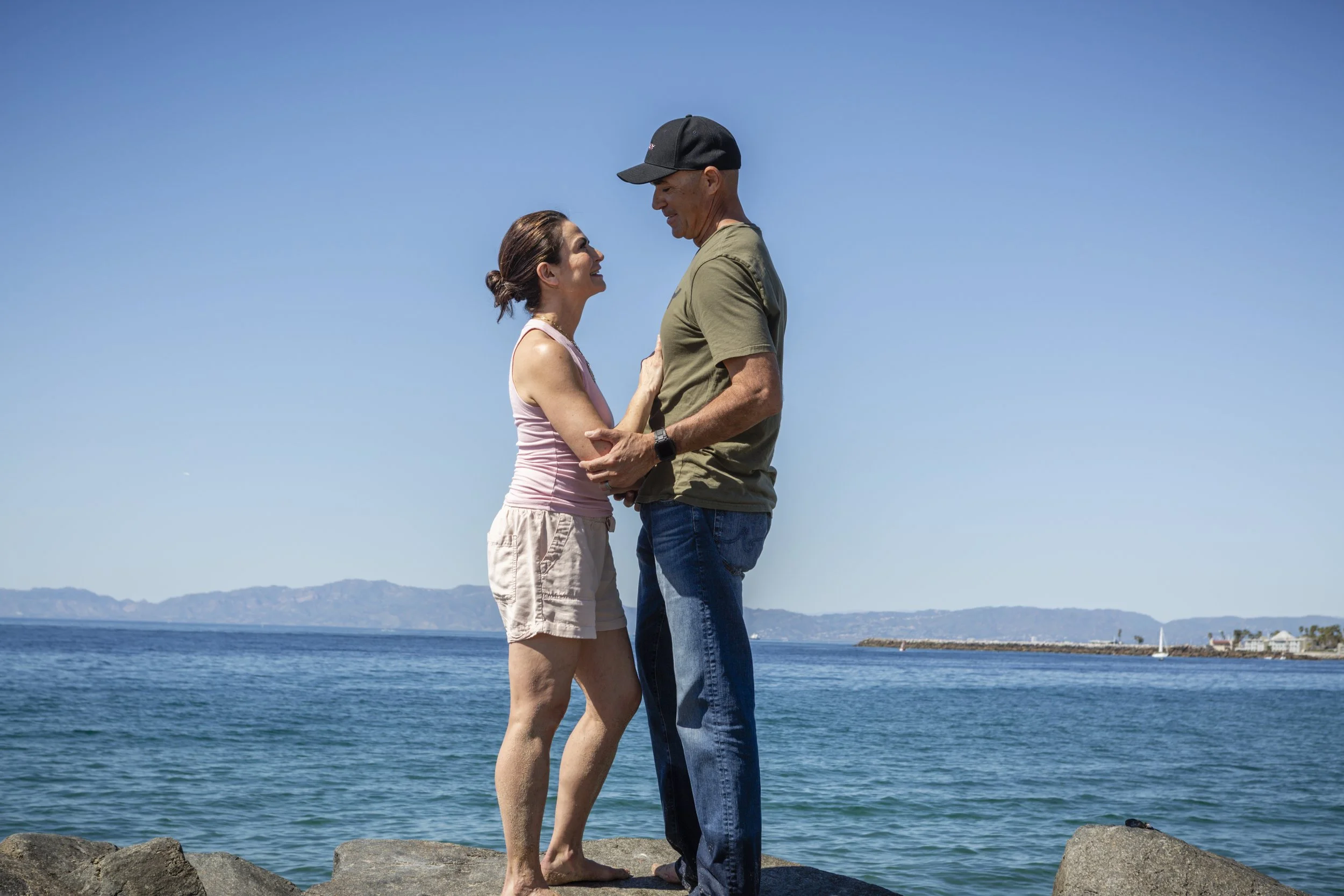 A couple stands on ocean rocks, enjoying the view and symbolizing healing, connection, and tranquility after infidelity through counseling.