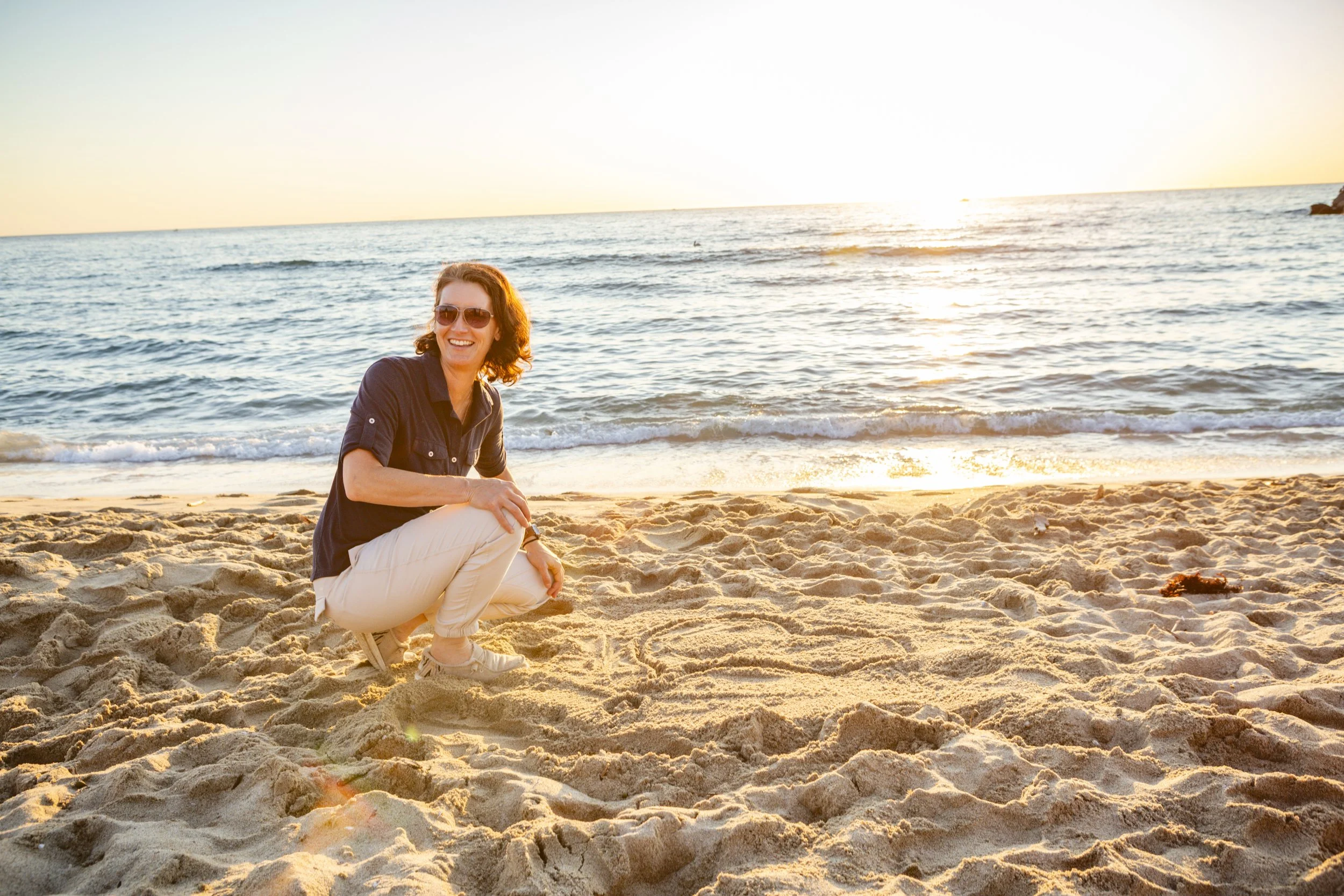 A woman with sunglasses and a dark shirt crouching on sandy beach during sunset with ocean waves in the background.