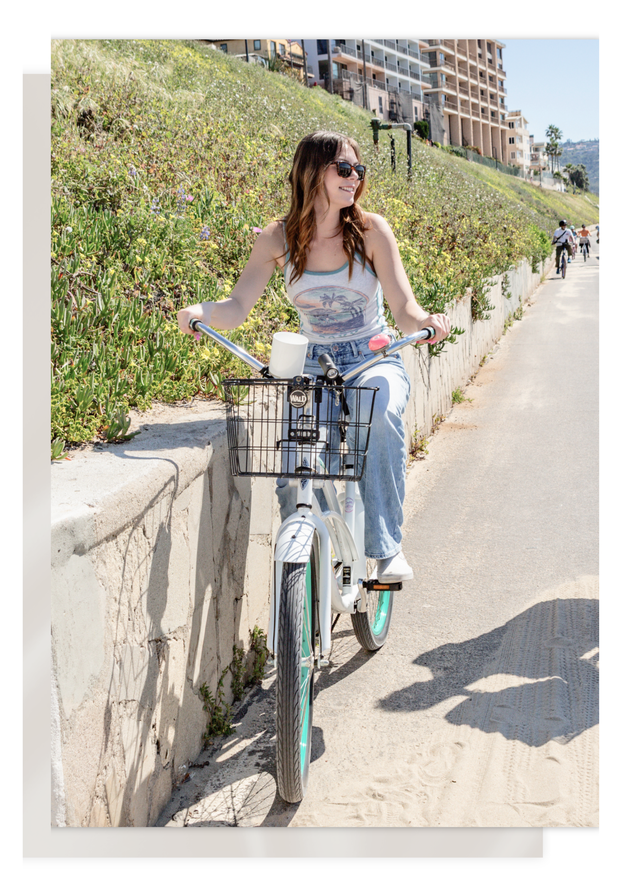 A smiling woman with long brown hair wearing sunglasses, riding a white bicycle along a sidewalk beside a garden with green plants and flowers.