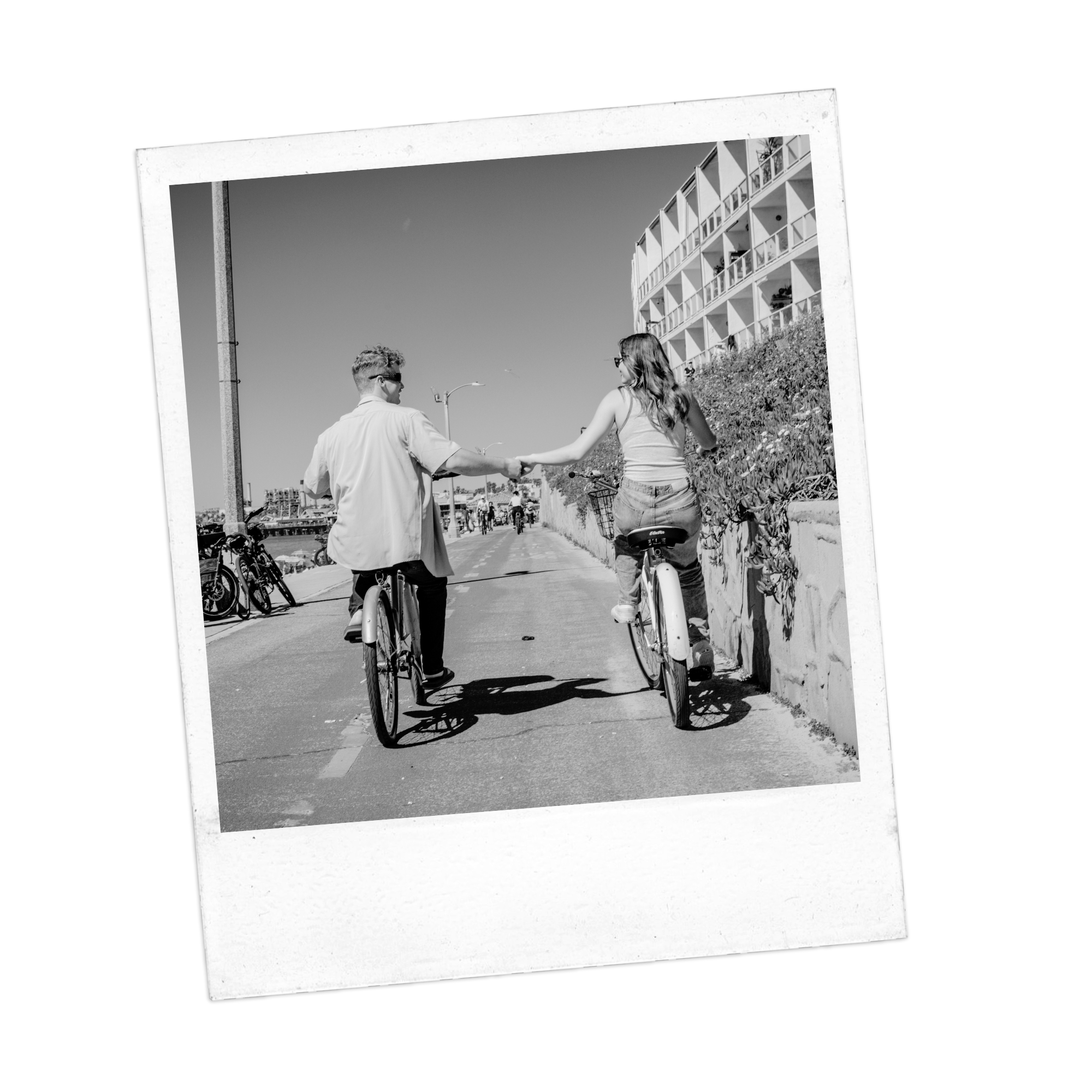 Black-and-white photo of a young couple on bikes holding hands and riding on a city sidewalk under a large framing structure, with apartment buildings and trees in the background.
