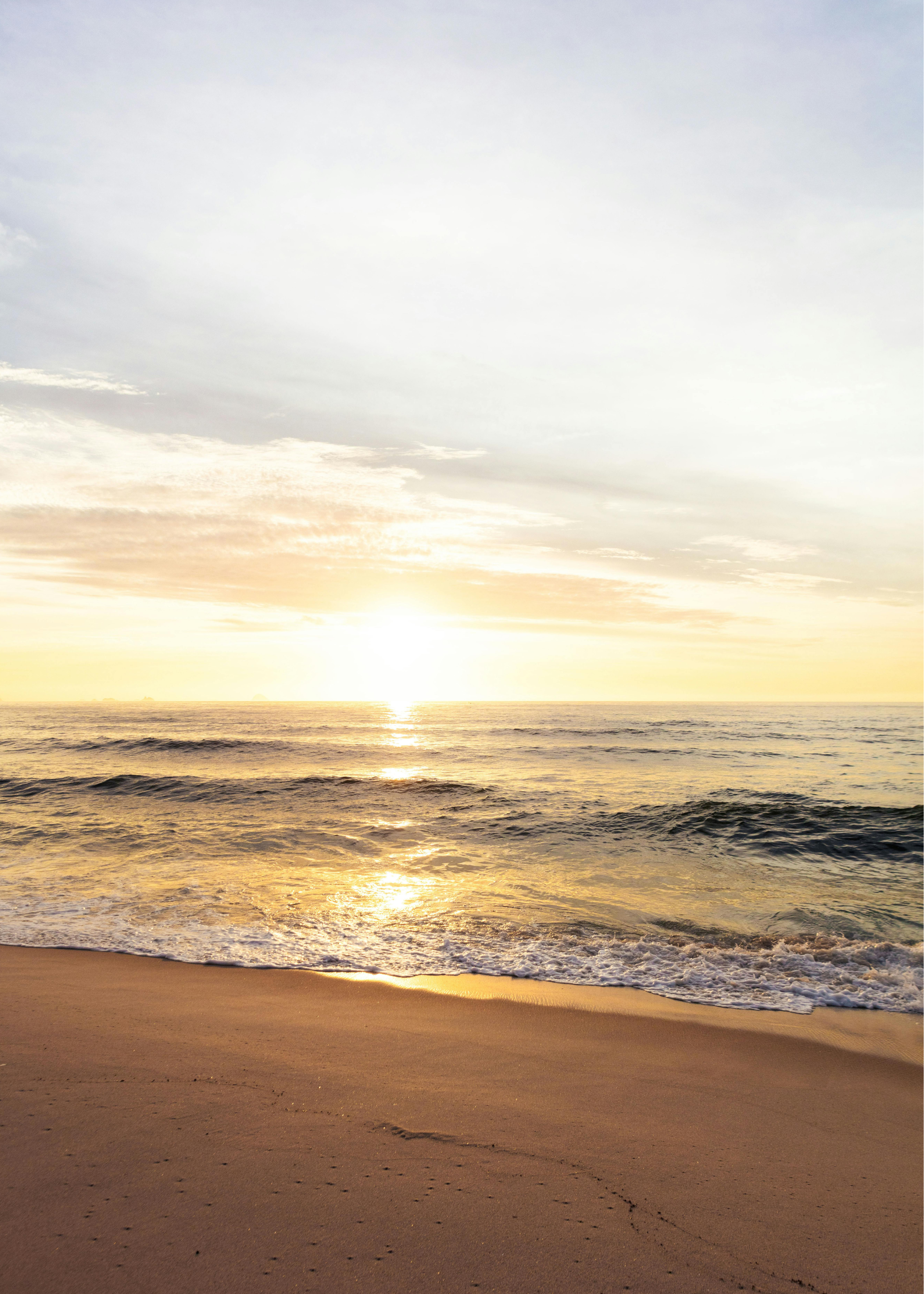 A peaceful beach scene during sunset or sunrise with a sandy shoreline, gentle waves, and a partly cloudy sky with the sun near the horizon.