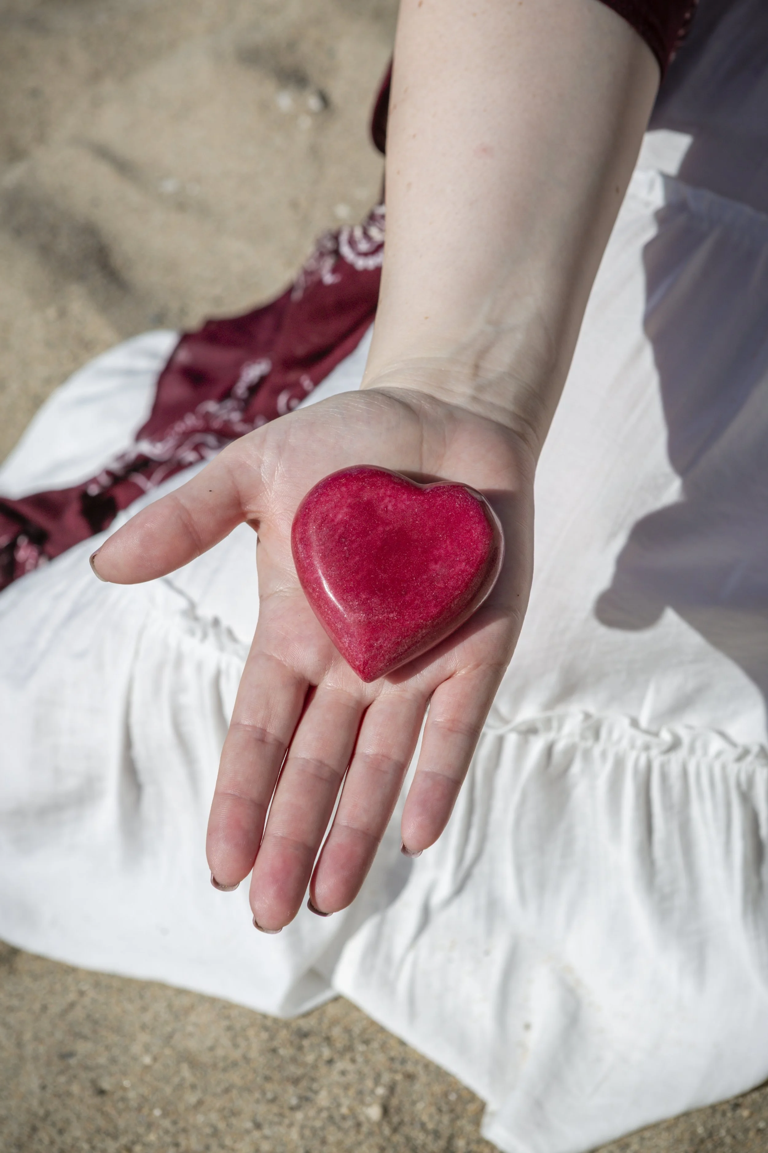 Person holding a red heart-shaped stone in their hand, wearing white pants and a maroon top, with a sandy ground background.
