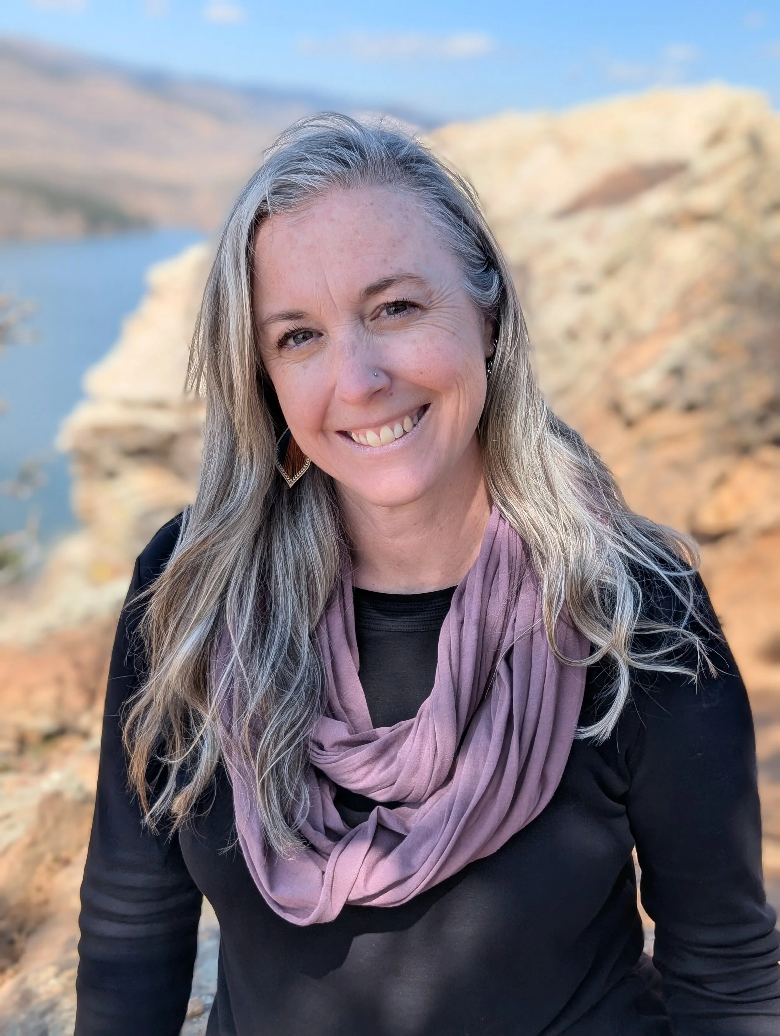 A woman smiling outdoors near a body of water with mountains in the background, wearing a black top and a pink scarf.