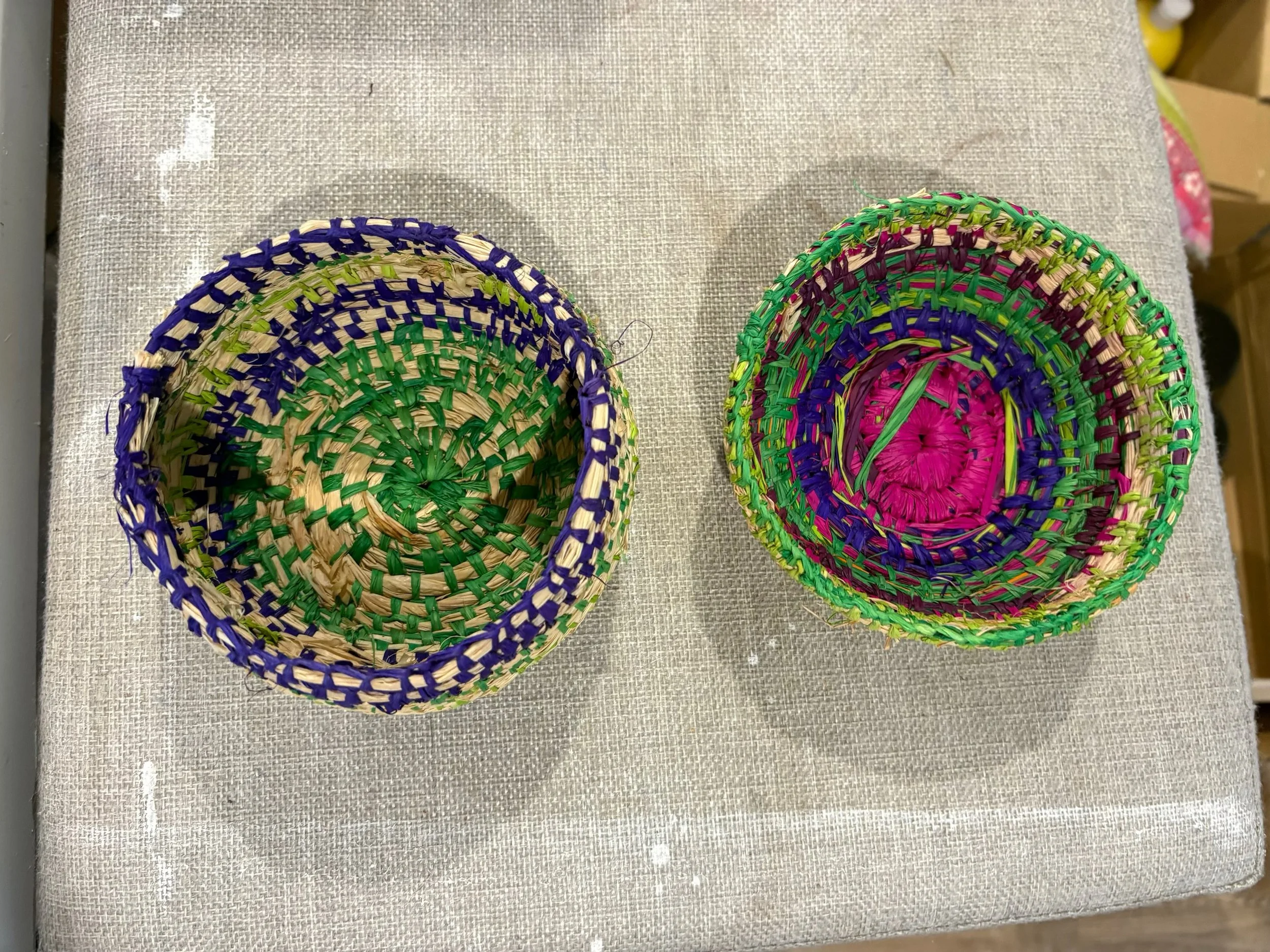 Two colorful woven baskets, one with purple and green details and one with pink, purple, and green details, placed on a light-colored fabric surface.