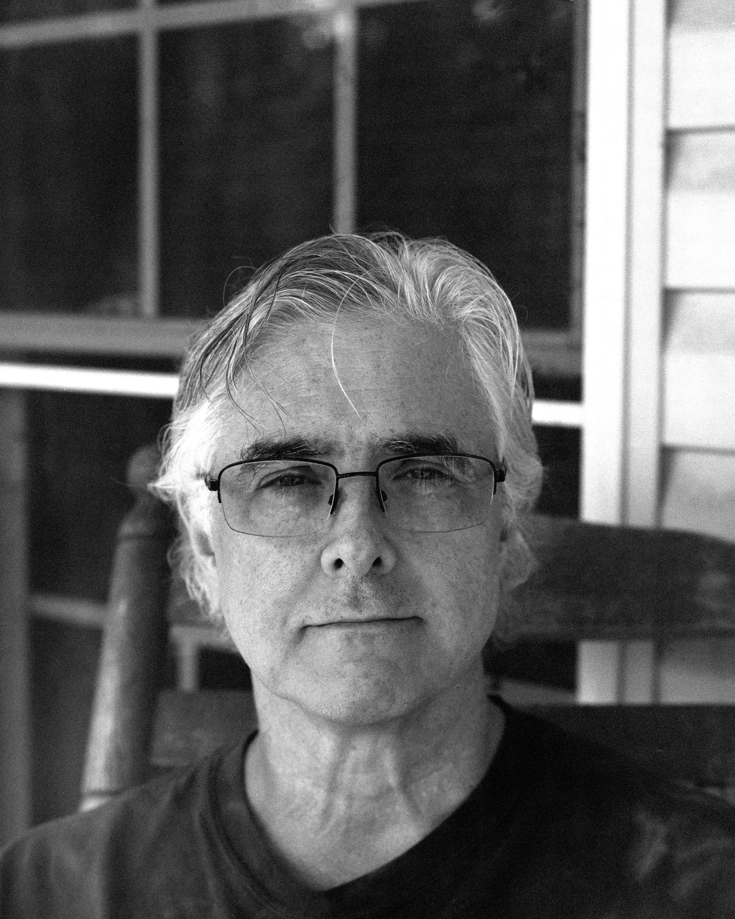 A black and white portrait of an older man with glasses and gray hair, sitting outdoors with a wooden chair and house siding in the background.