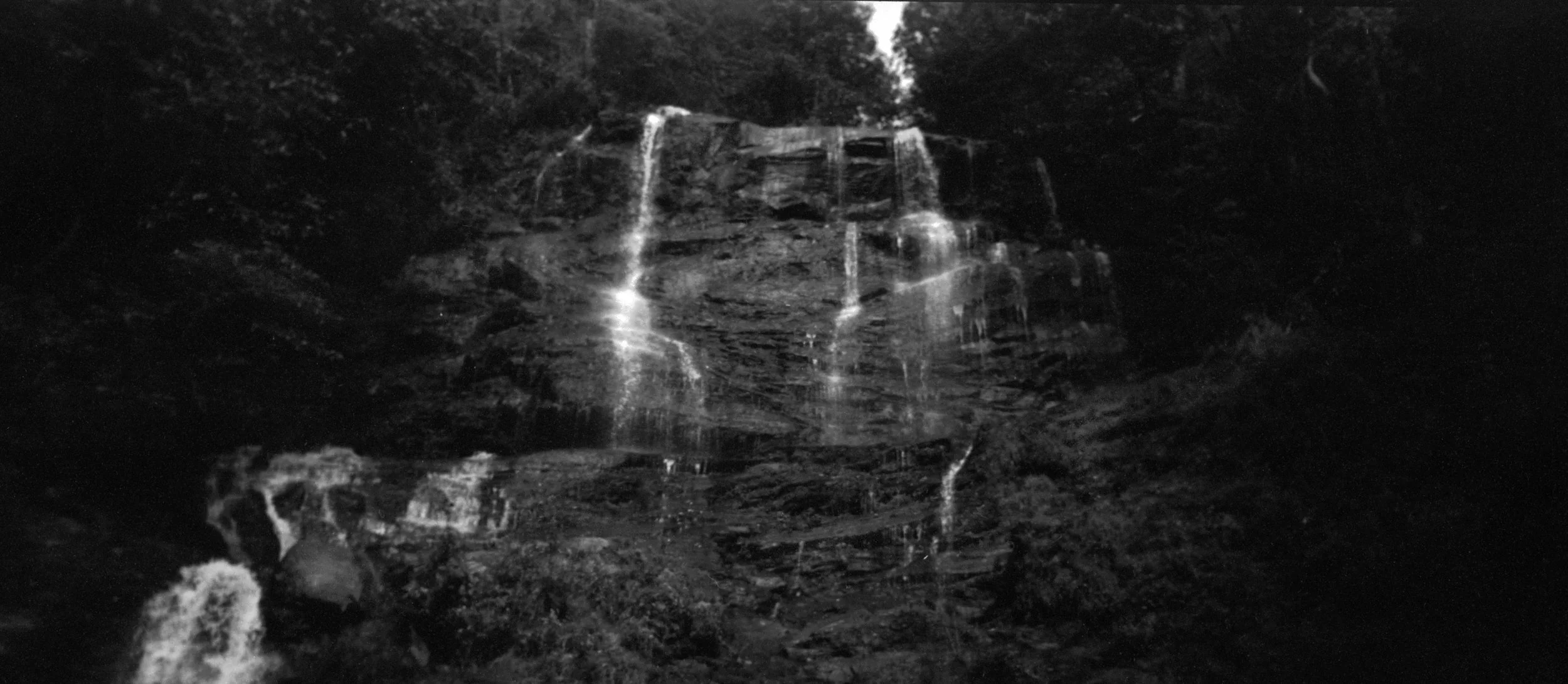 Black and white photo of a waterfall cascading down a rocky cliff surrounded by dense trees.