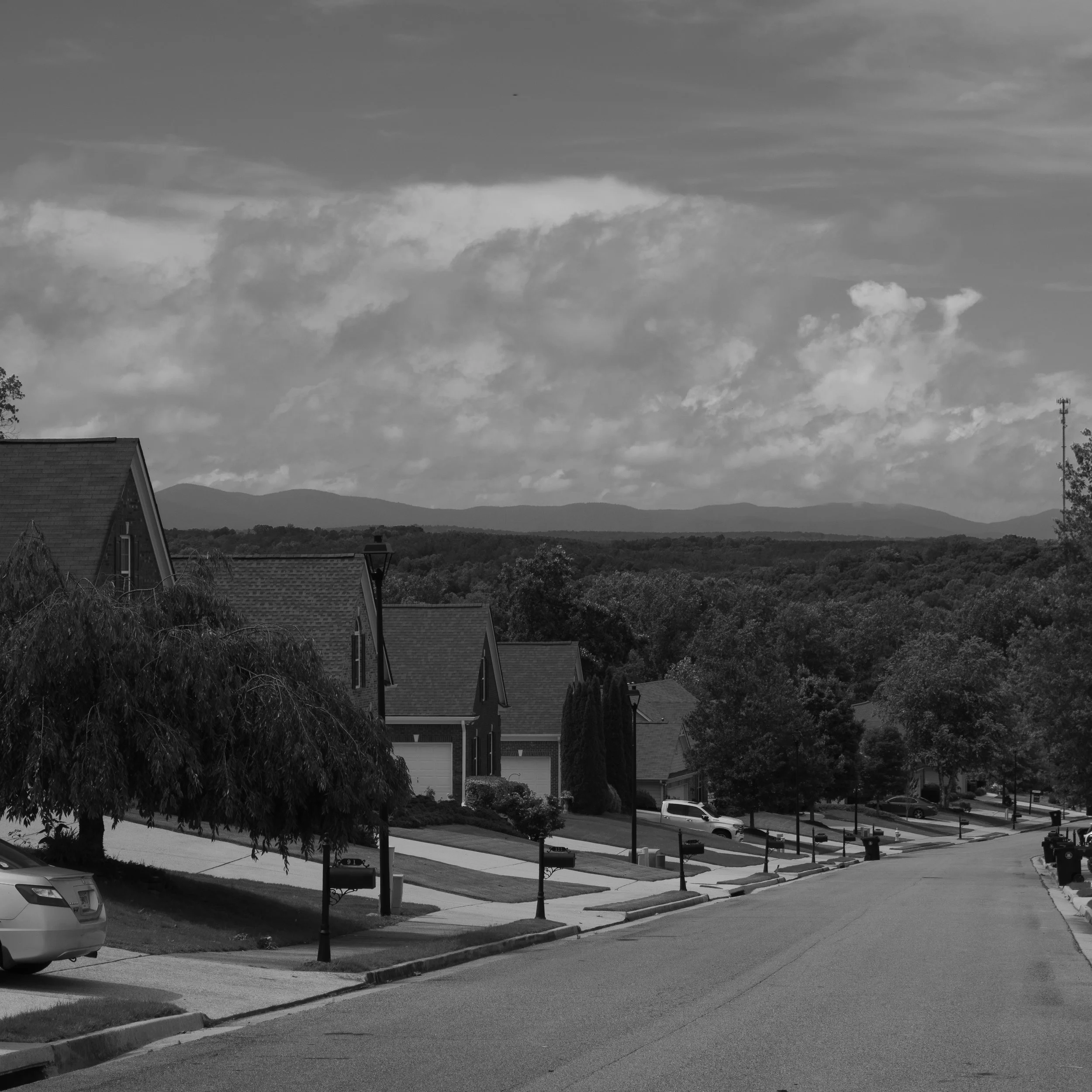 Residential neighborhood street with houses, parked cars, trees, street lamps, and mountains in the background under a partly cloudy sky.