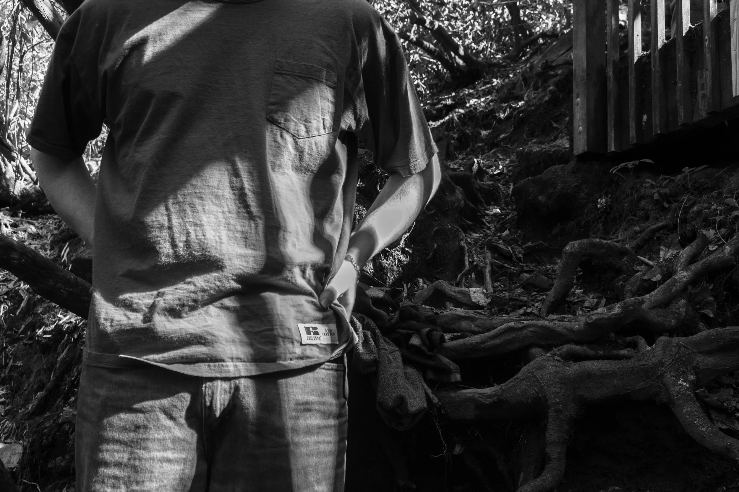 Person wearing a t-shirt and jeans standing outdoors in a wooded area with a dirt path, roots, and a fence in the background.