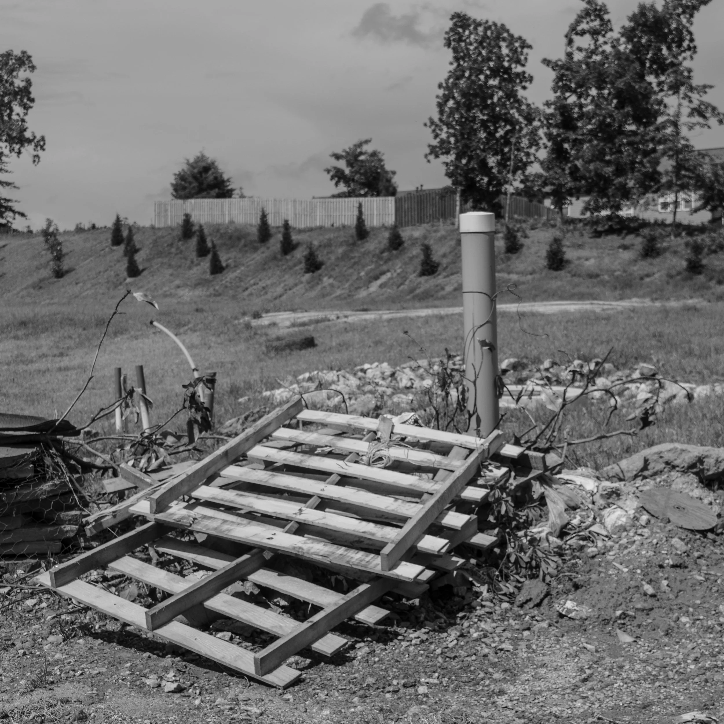 A fallen wooden pallet lying on the ground beside a broken pipe and some discarded debris in an outdoor area with grass, trees, and fencing in the background.