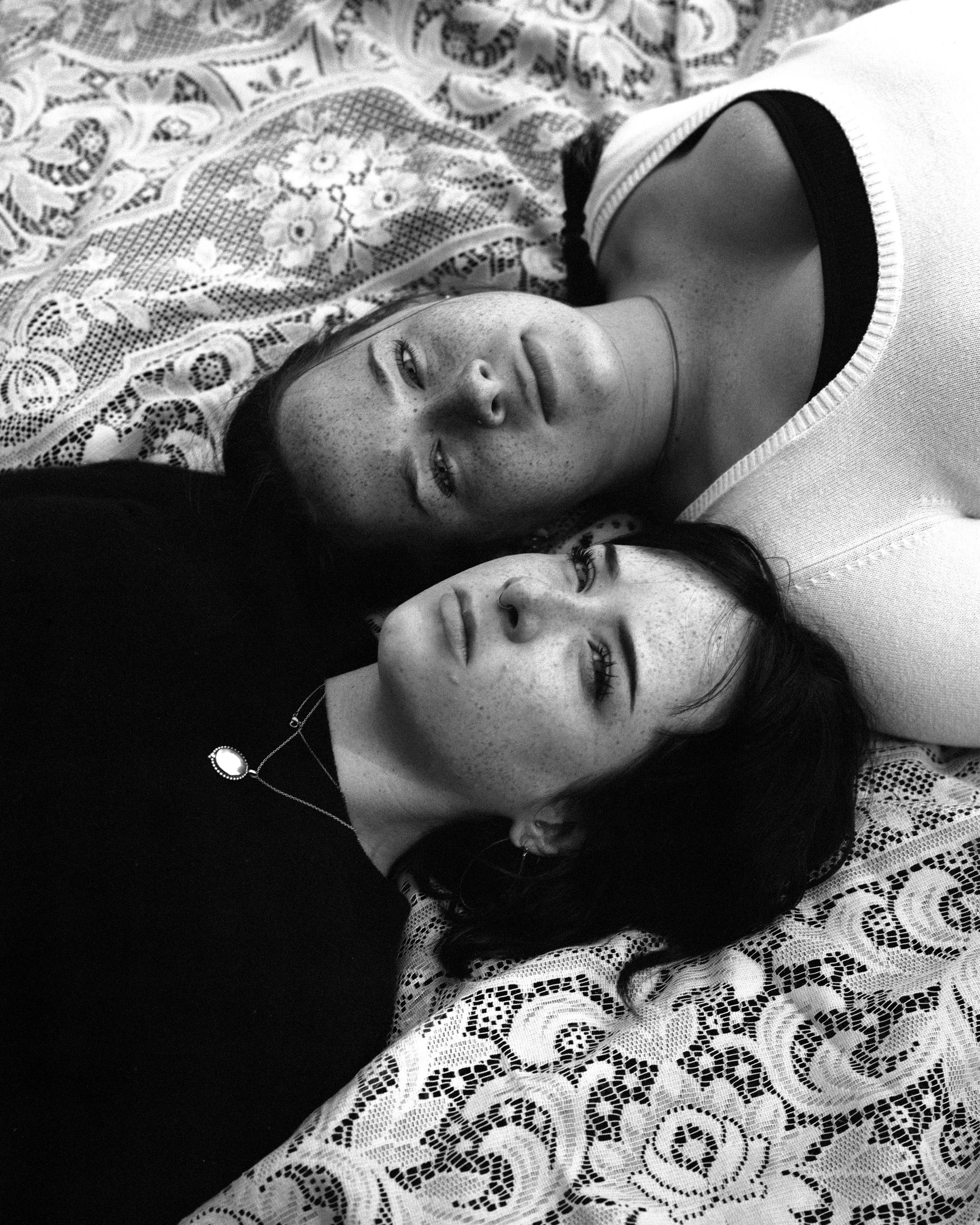 Two women lying on a lace patterned fabric, facing opposite directions, with their heads close together. The woman on top has light skin, freckles, and long, wavy hair, wearing a white shirt. The woman below has darker skin, freckles, dark curly hair, and a necklace with a white pendant.