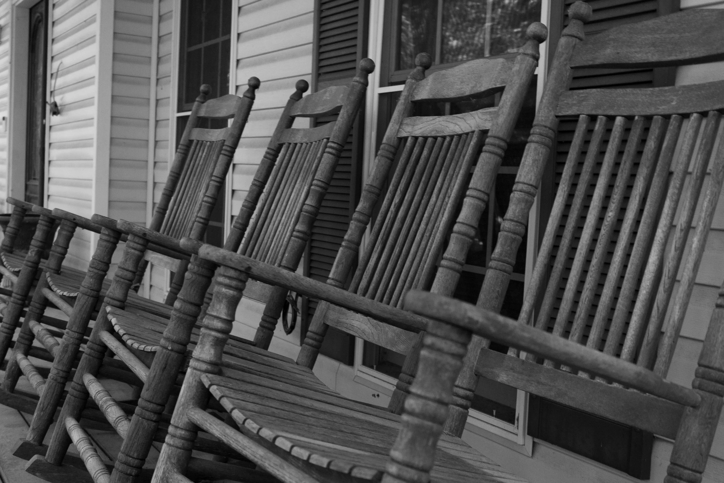 Three wooden rocking chairs lined up on a porch in front of a house with striped siding and windows.