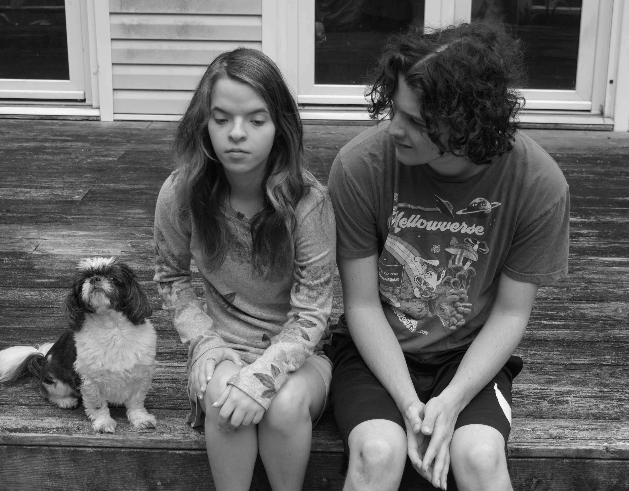 A black and white photo of a girl and a teenage boy sitting on a wooden deck with a small dog next to them, near sliding glass doors.