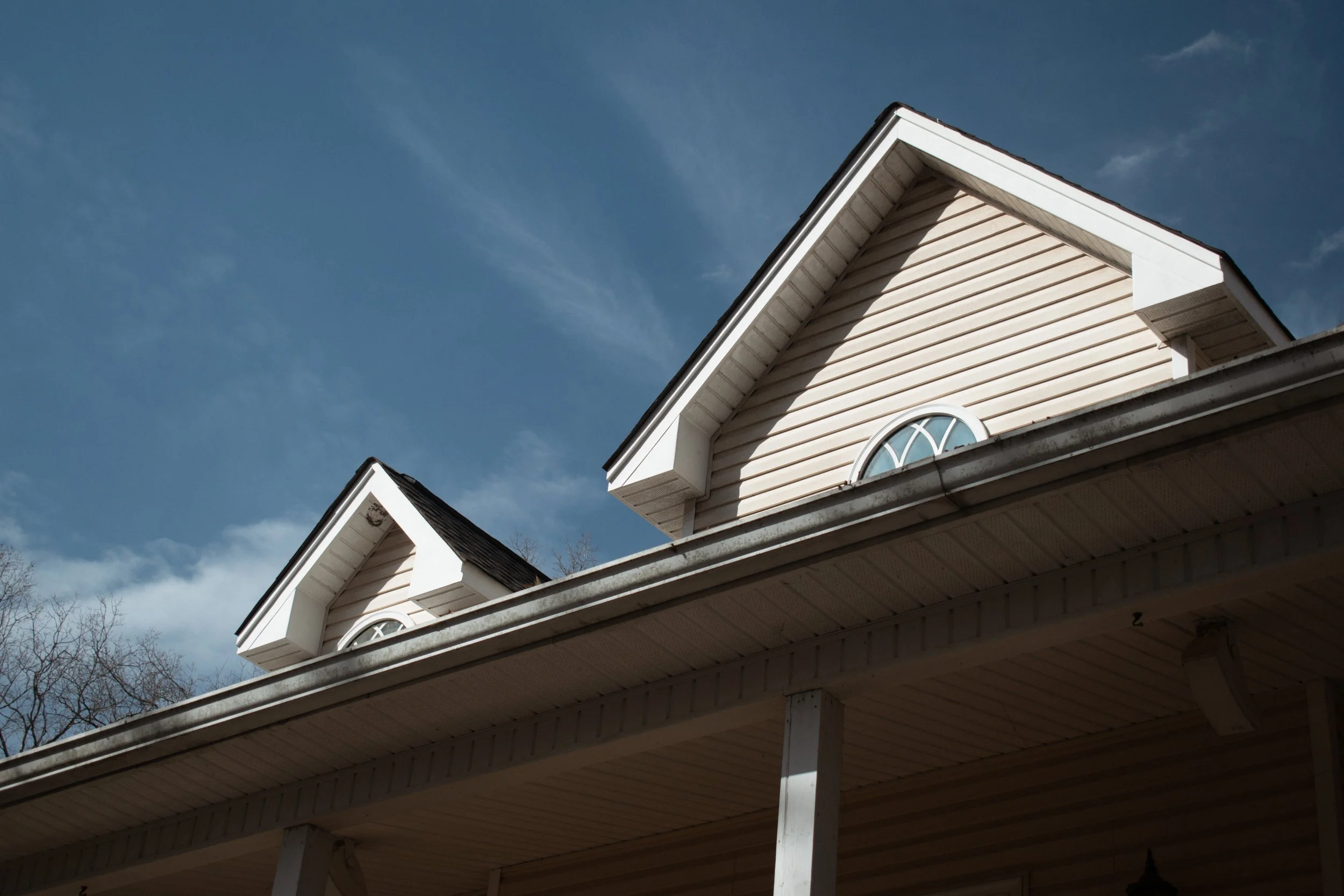 Close-up of the upper part of a house with beige siding and a gable roof, featuring small arched windows and a blue sky with some clouds in the background.