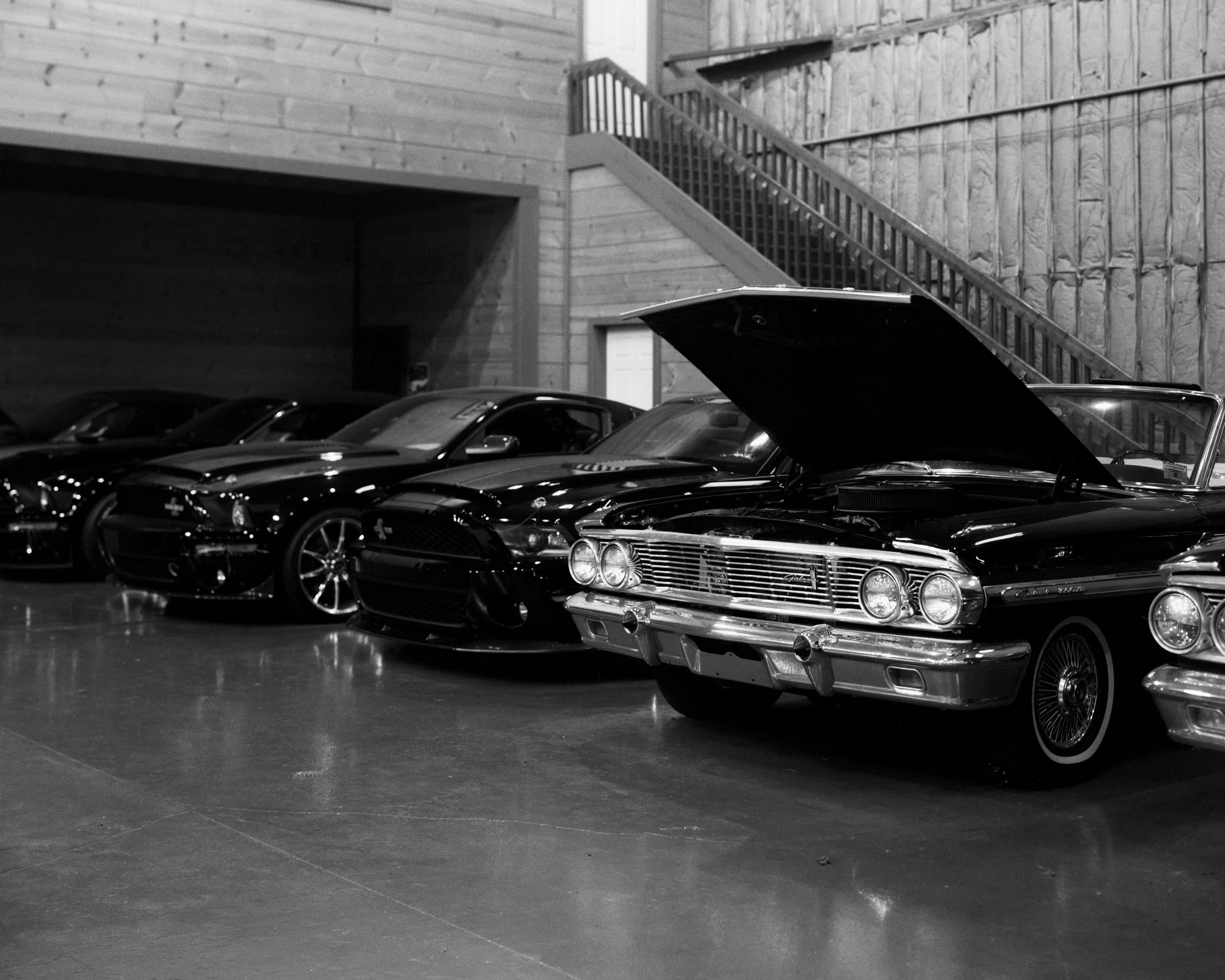 Black and white photograph of a row of classic cars inside a garage or showroom, with one car in the foreground having its hood open.
