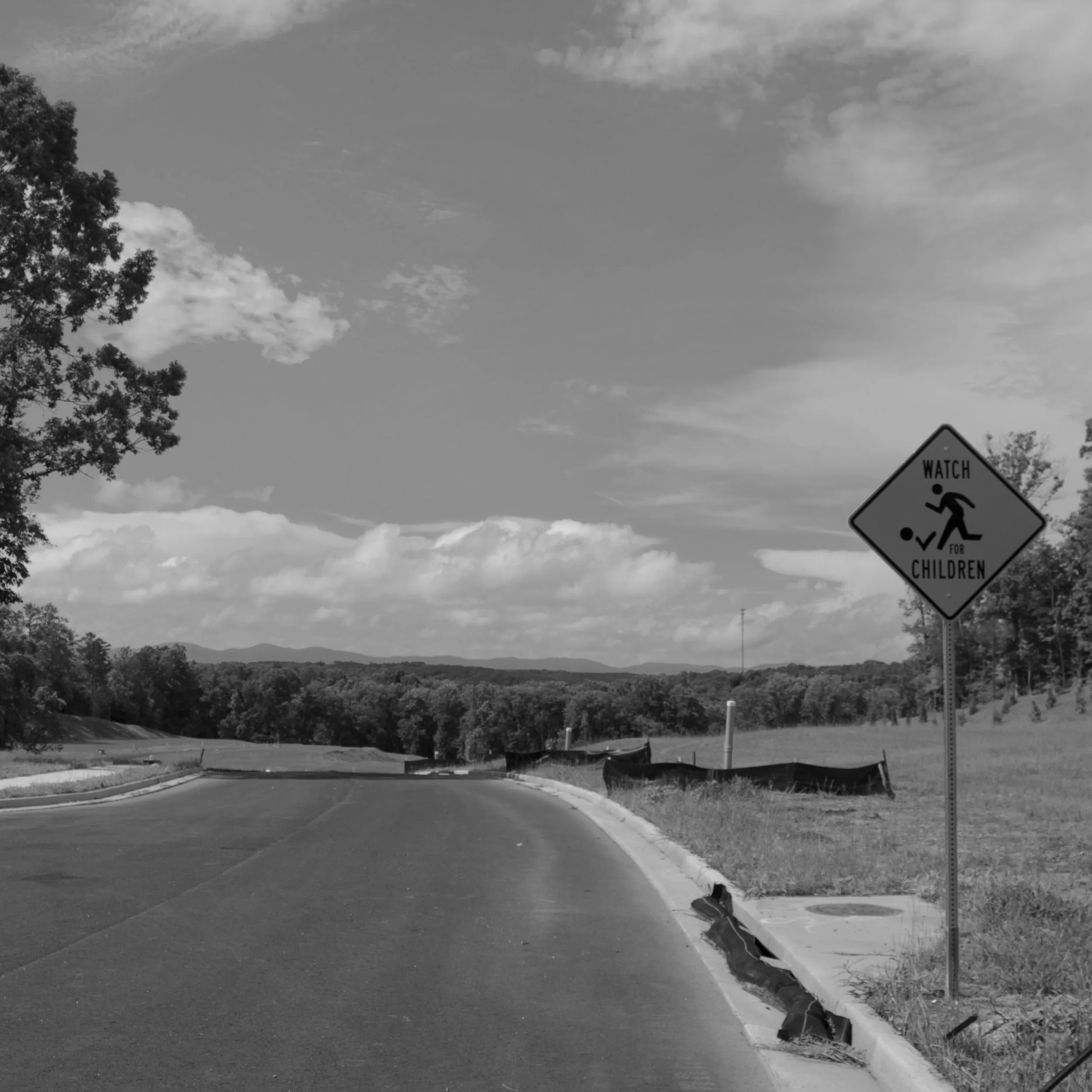 Road leading into a rural area with a sign that reads "Watch for Children" next to the sidewalk, with trees and rolling hills in the background under a partly cloudy sky.