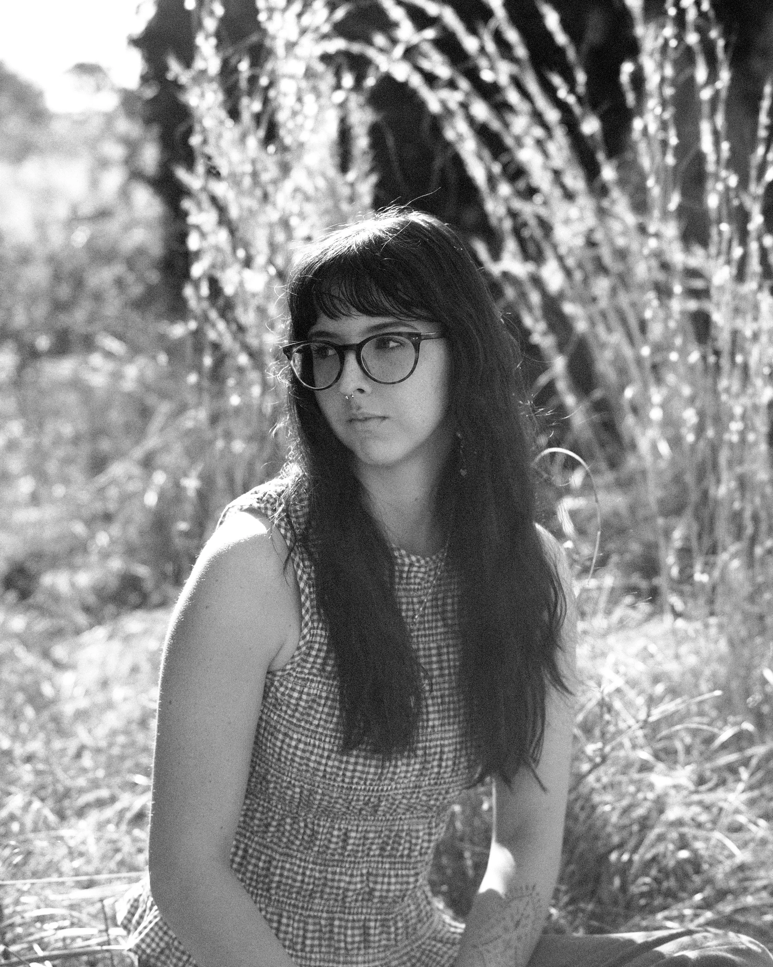 Black and white photo of a young woman with glasses and dark hair sitting outdoors surrounded by plants and trees.