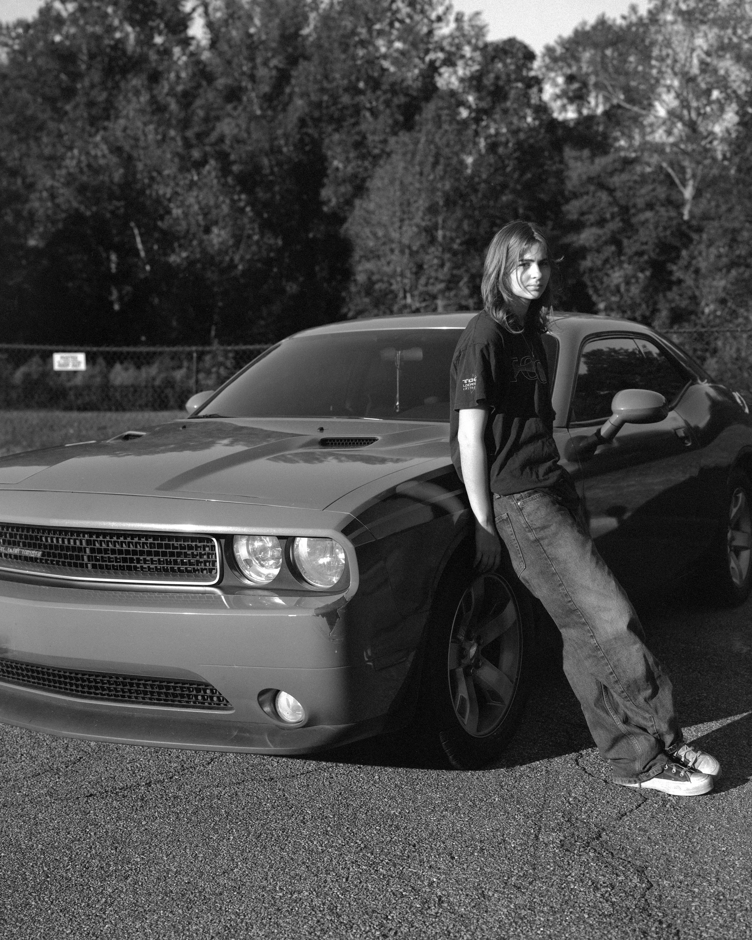 A young woman with shoulder-length hair in casual clothing leaning against a modern, sleek sports car parked in an outdoor lot with trees in the background.