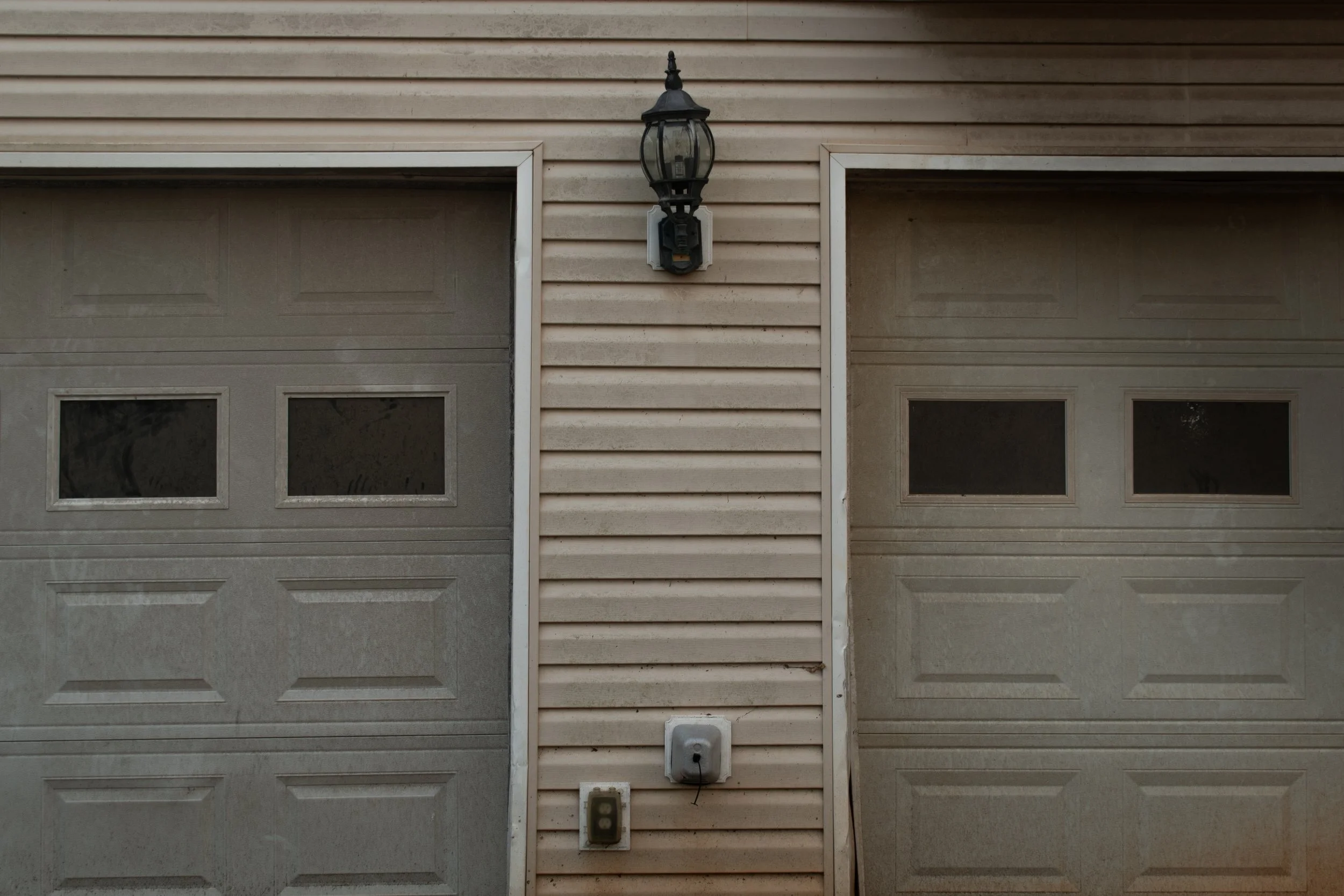 Double garage doors on either side of a beige siding wall with three small windows on each garage door, an outdoor lantern-style light fixture above, and an electrical outlet with a cover below.