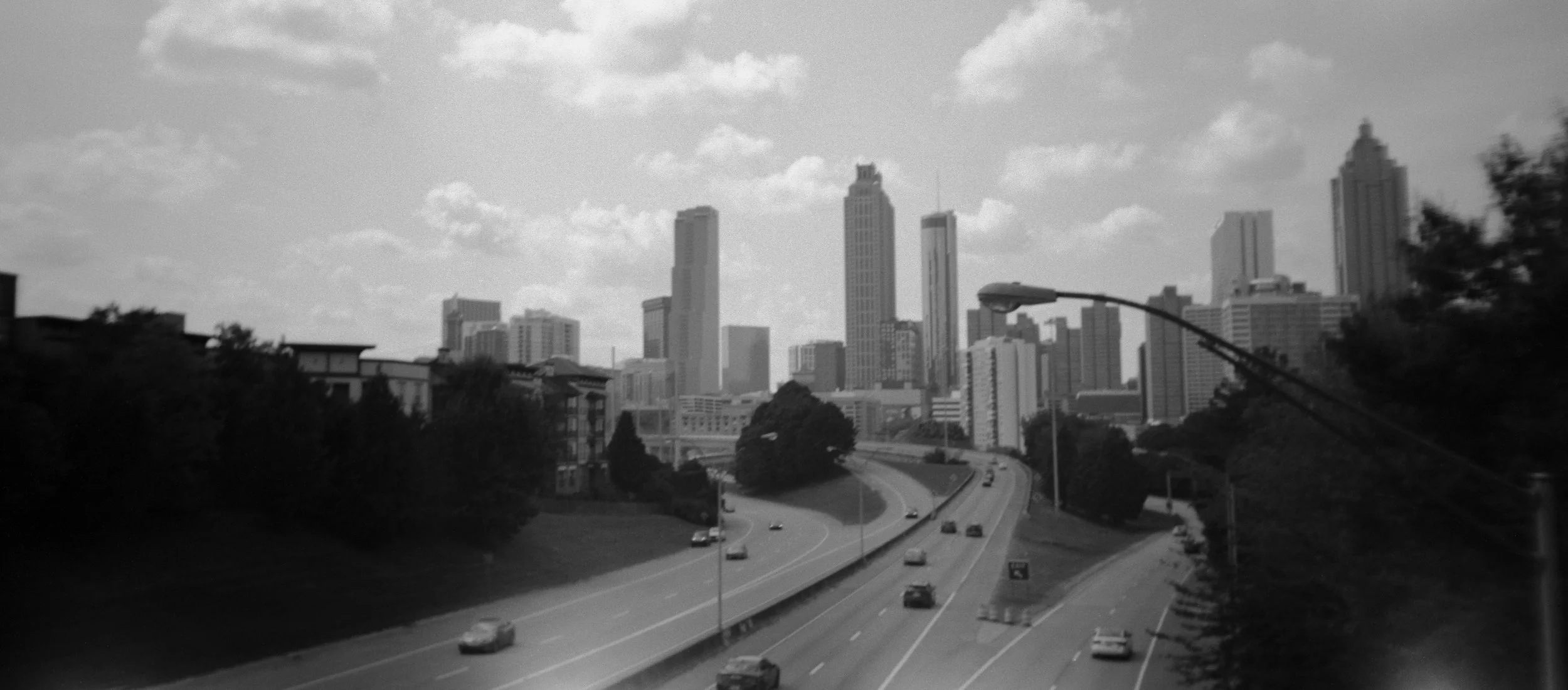 Black and white photo of a city skyline with tall buildings and skyscrapers, viewed from a highway with moving cars and trees on the sides. A street lamp is visible in the foreground.