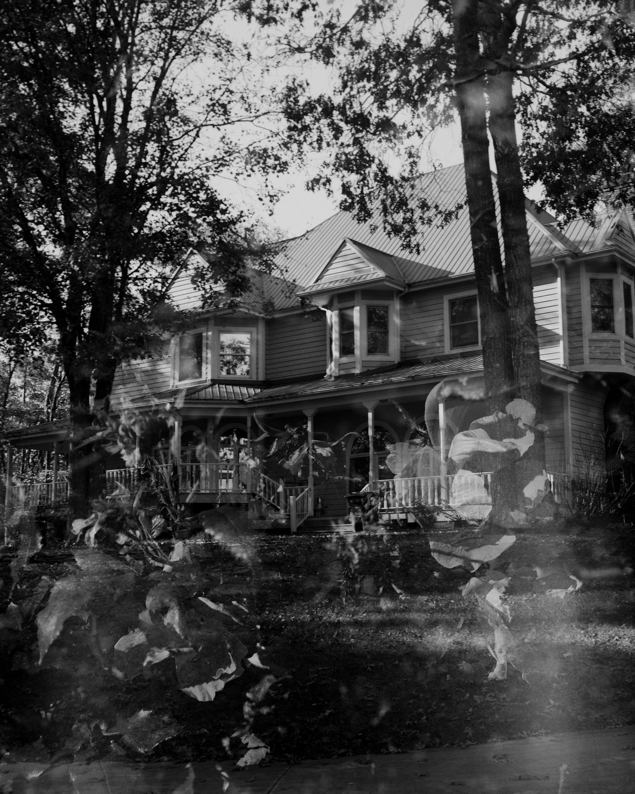 A black and white photograph of a house surrounded by trees, with reflections of leaves and a person in a hat on a window or glass surface in the foreground.