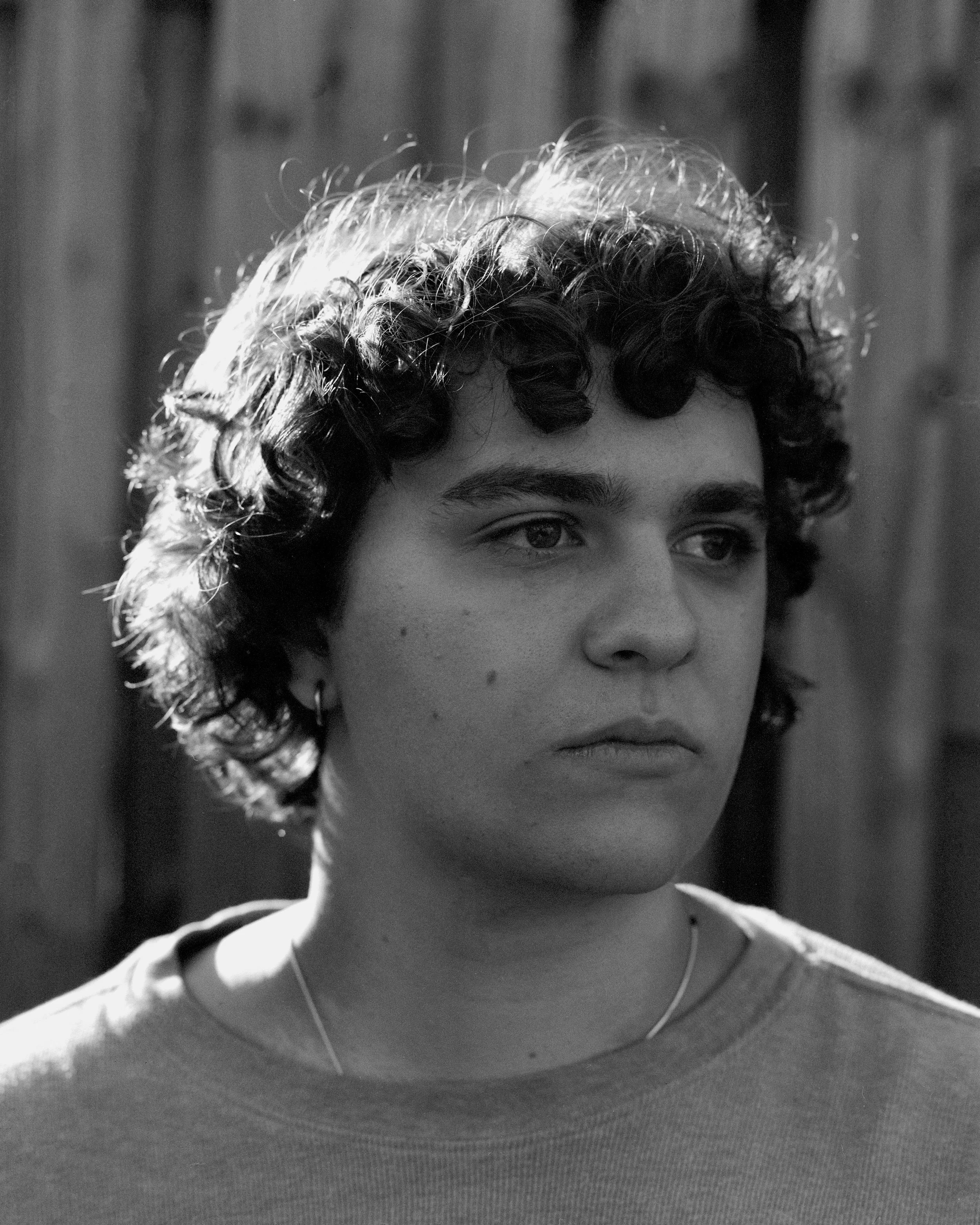 Black and white close-up portrait of a young woman with curly hair, wearing a t-shirt and a necklace, standing outdoors with a blurred wooden fence background.
