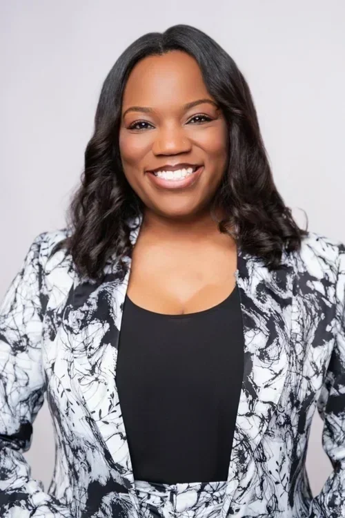 A smiling African American woman with shoulder-length wavy black hair, wearing a black top and a black and white patterned blazer, against a light gray background.