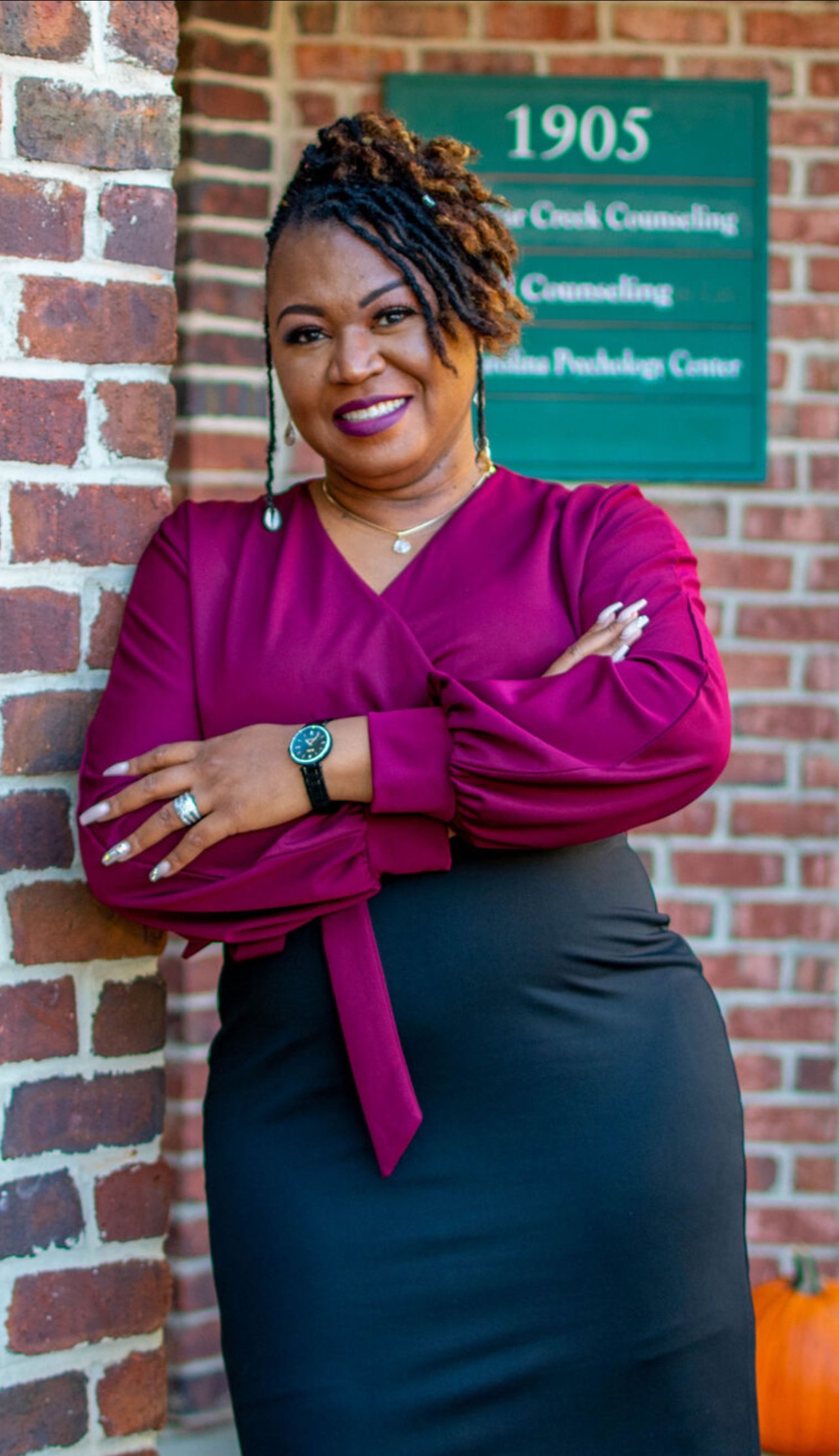 A woman with styled dreadlocks wearing a purple blouse and black skirt, smiling and standing outdoors against a brick wall with a green sign behind her that reads '1905.'
