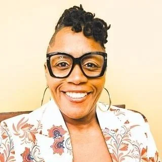 A woman with short curly hair, wearing glasses and large hoop earrings, smiling in front of a plain yellow wall, dressed in a patterned shirt.