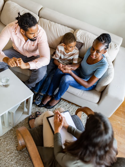 A family therapy session with a woman therapist taking notes while a family of three, including a man, a woman, and a young boy, sit on a beige couch and talk to her.