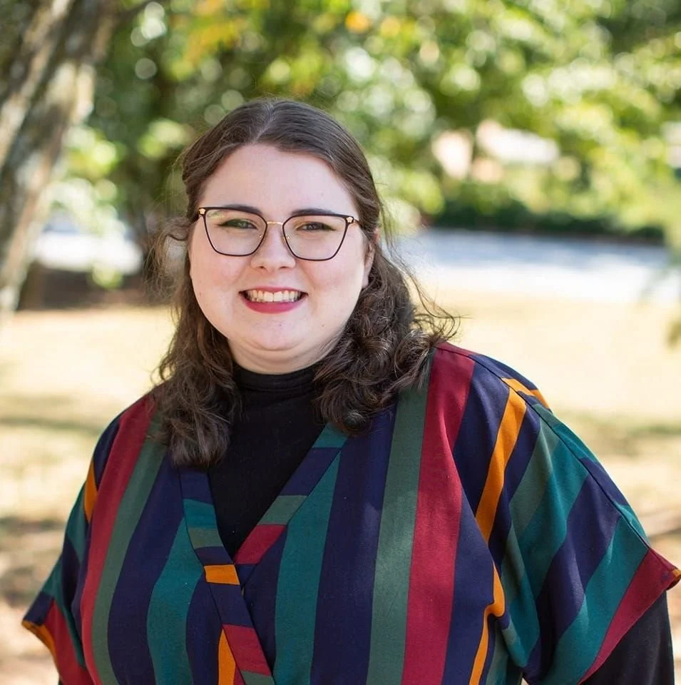 A woman standing outdoors with trees and a fence in the background, wearing glasses and a colorful striped jacket while smiling at the camera.