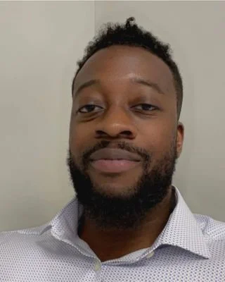 Close-up of a man with short curly hair and a beard, wearing a collared shirt, standing against a plain background.