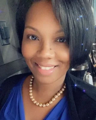 A smiling woman with short black hair wearing a blue top and a pearl necklace.