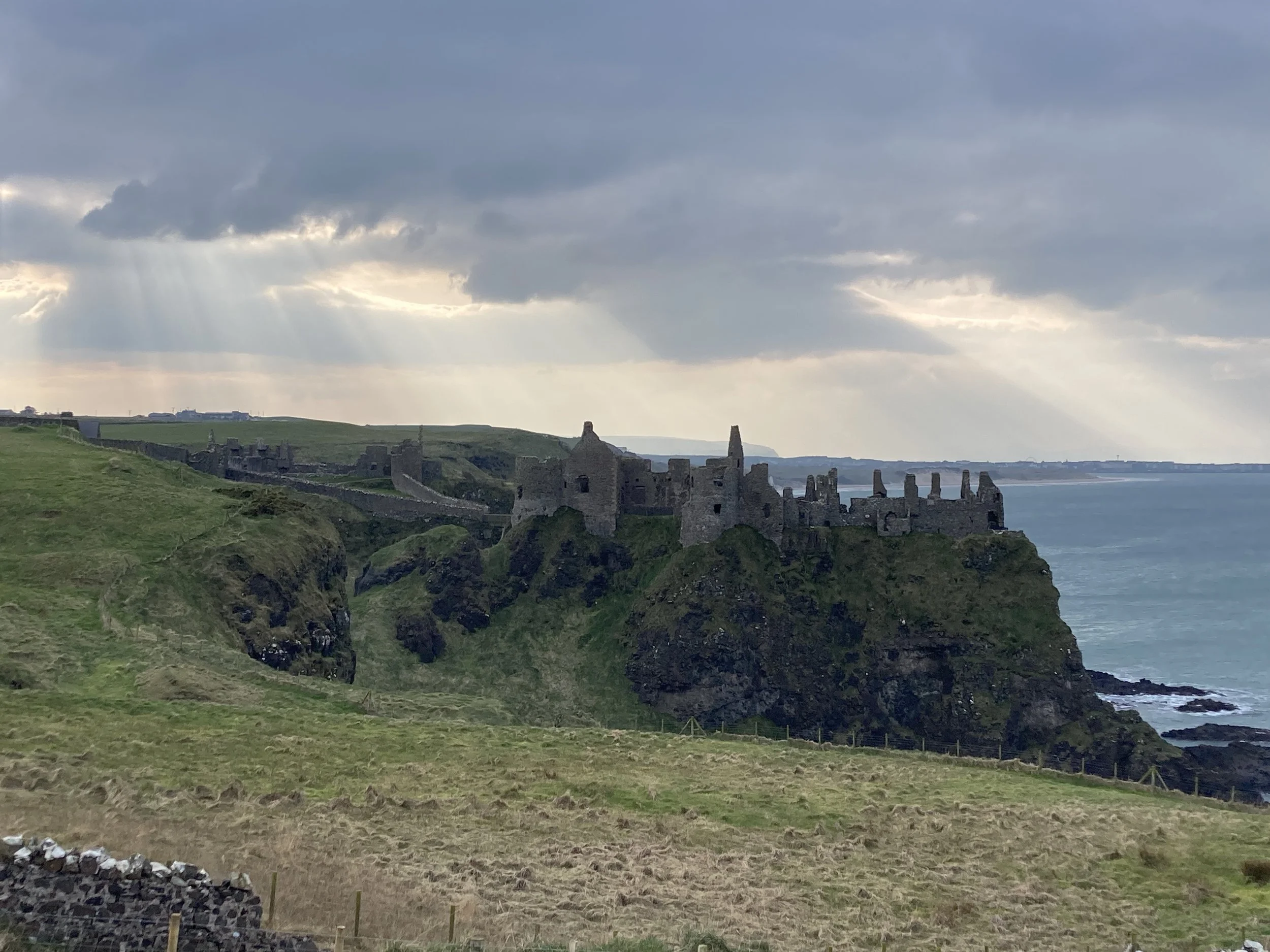 Ruin of Dunluce Castle, Bushmills, County Antrim, Northern Ireland, atop a green cliff overlooking the ocean under a cloudy sky with rays of sunlight breaking through.