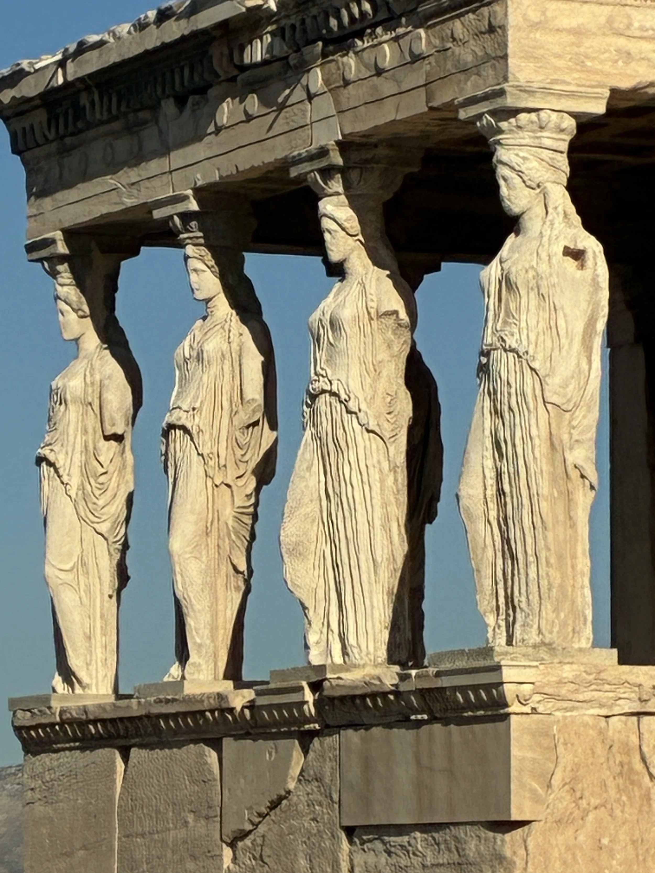 Close-up of the Porch of the Caryatids on the Acropolis of Athens, carved from marble, with worn detailing, standing in a row on a stone platform under a clear blue sky.