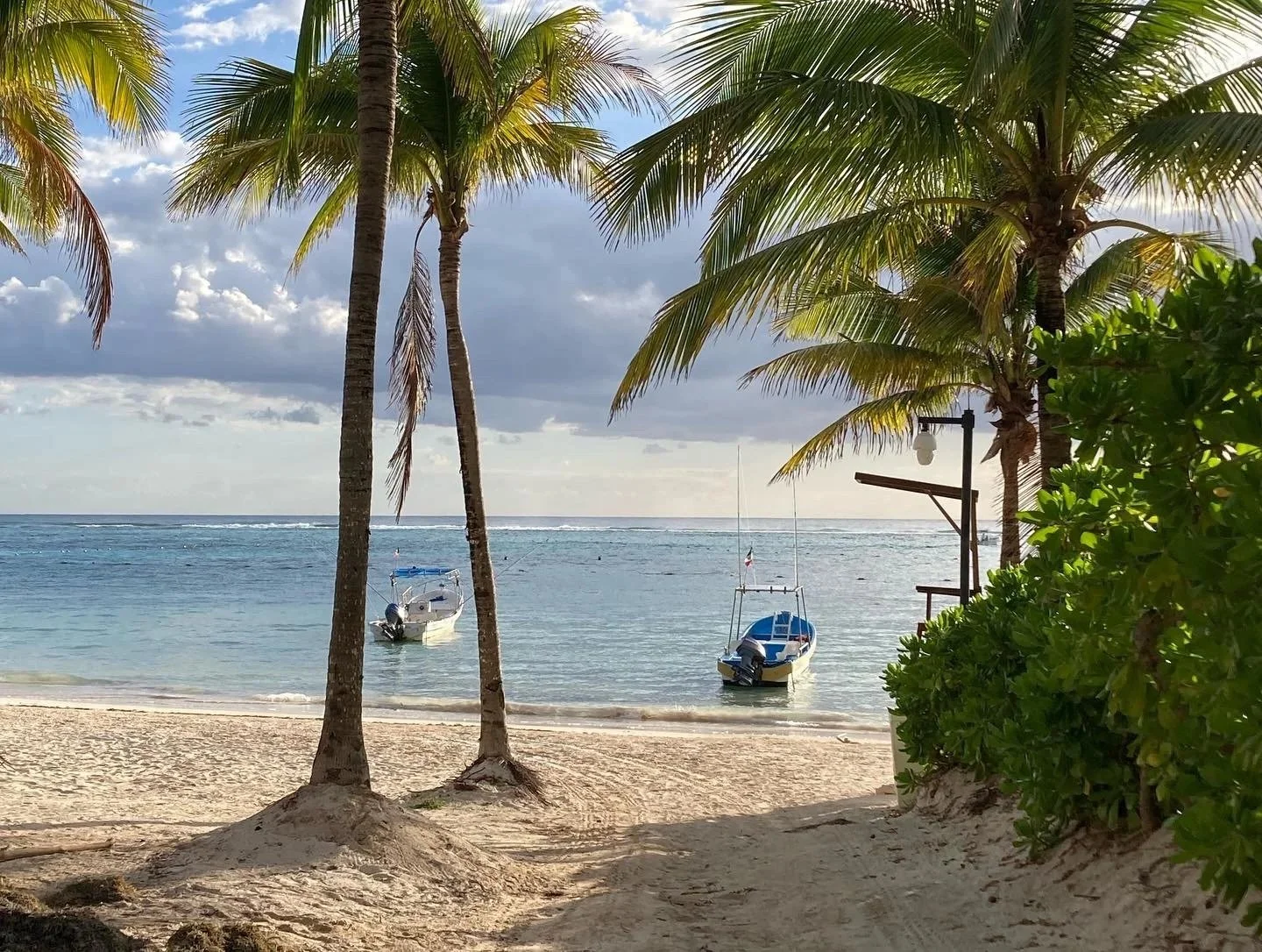 A tropical beach in Akumal, Mexico, with sandy shore, two palm trees, and lush green bushes. Two small boats float in calm water, and a cloudy sky is visible in the background.
