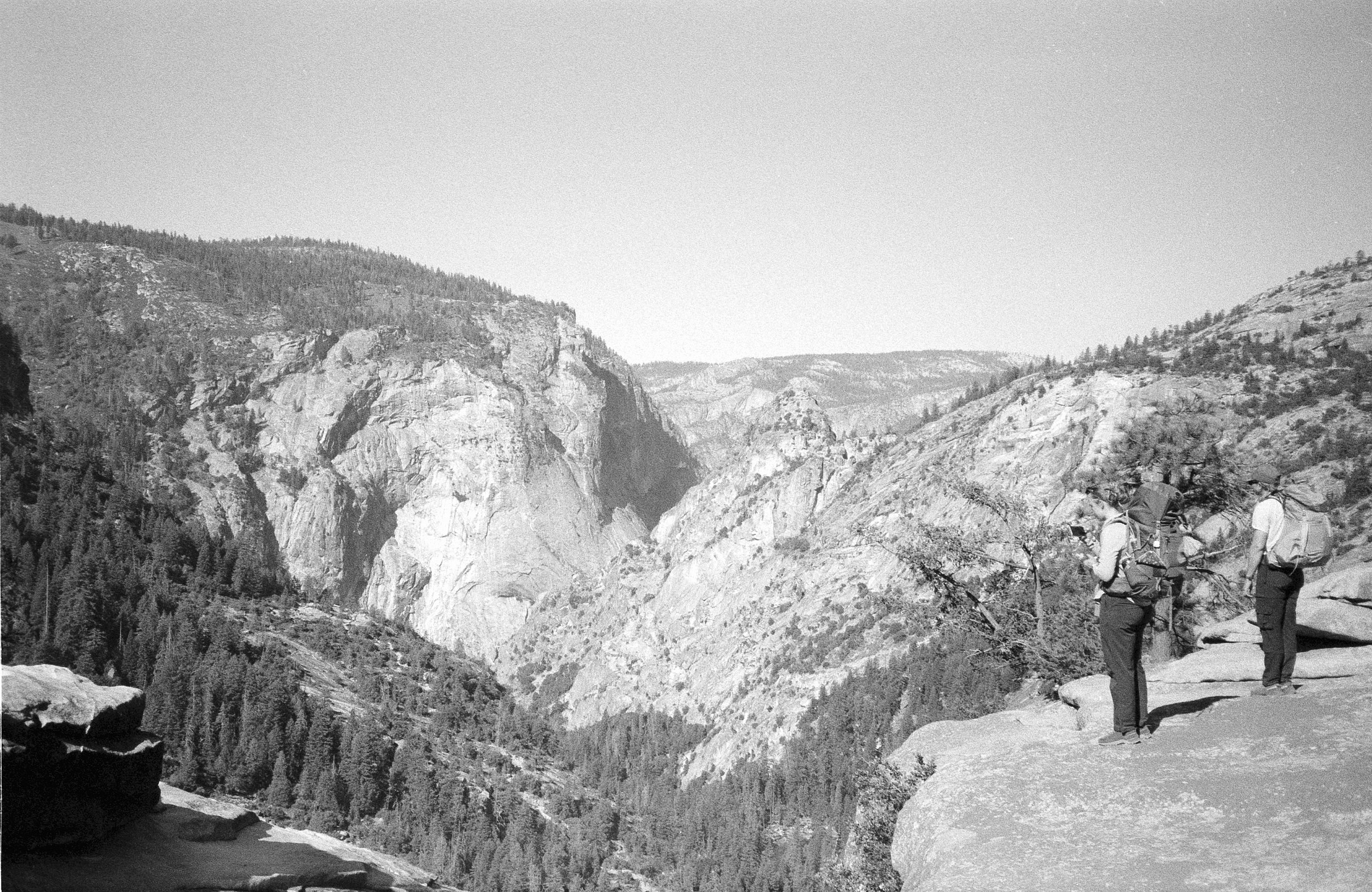 Two hikers with backpacks standing on a rocky outcrop overlooking a canyon with steep cliffs and dense forested areas.