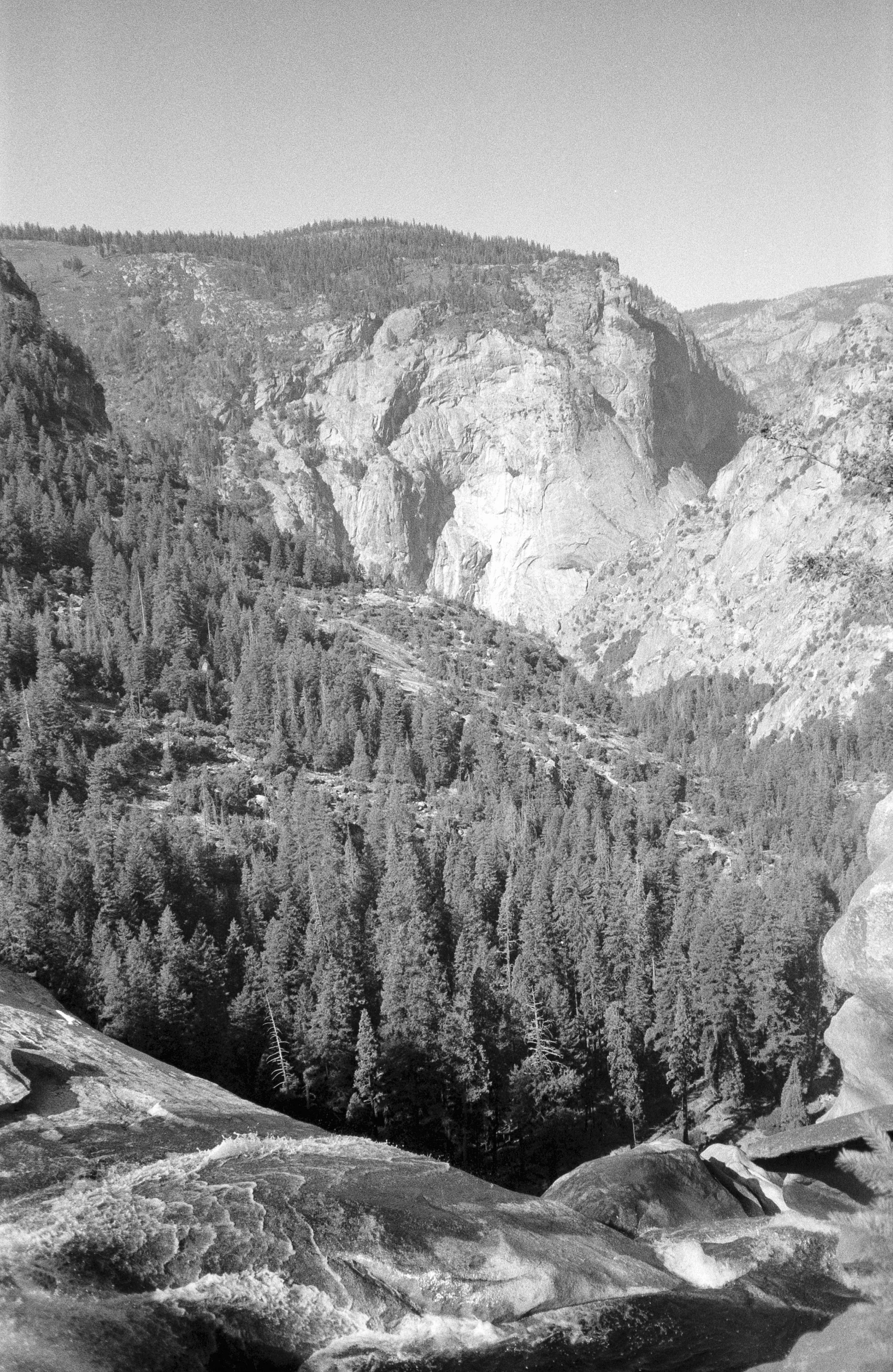 Black and white photograph of a mountainous landscape with cliffs and rugged terrain, dense forest of coniferous trees, and a rocky foreground.