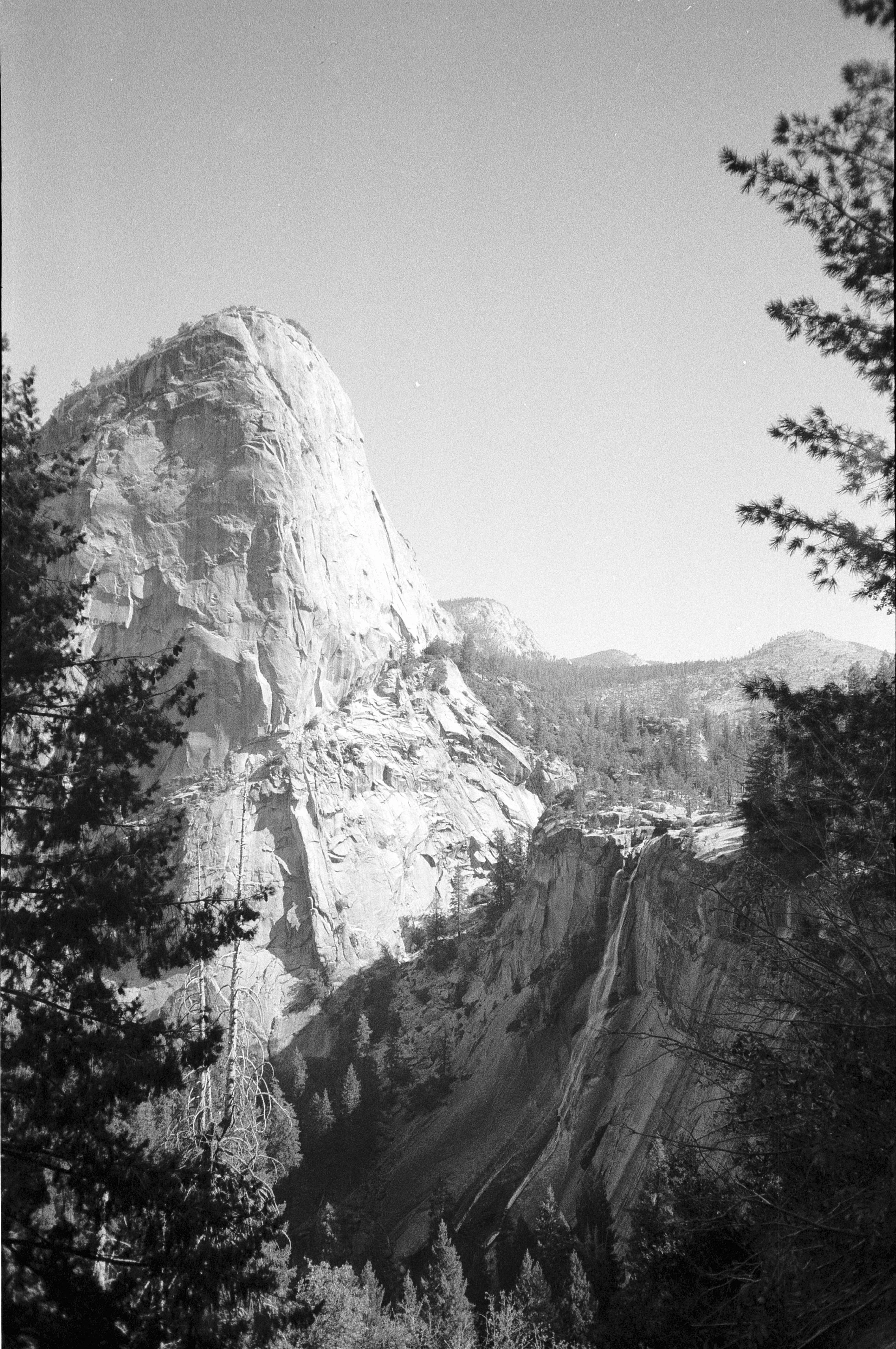 Black and white photograph of a mountain landscape with a large, rounded rock formation on the left, surrounded by trees, and a waterfall on the right.