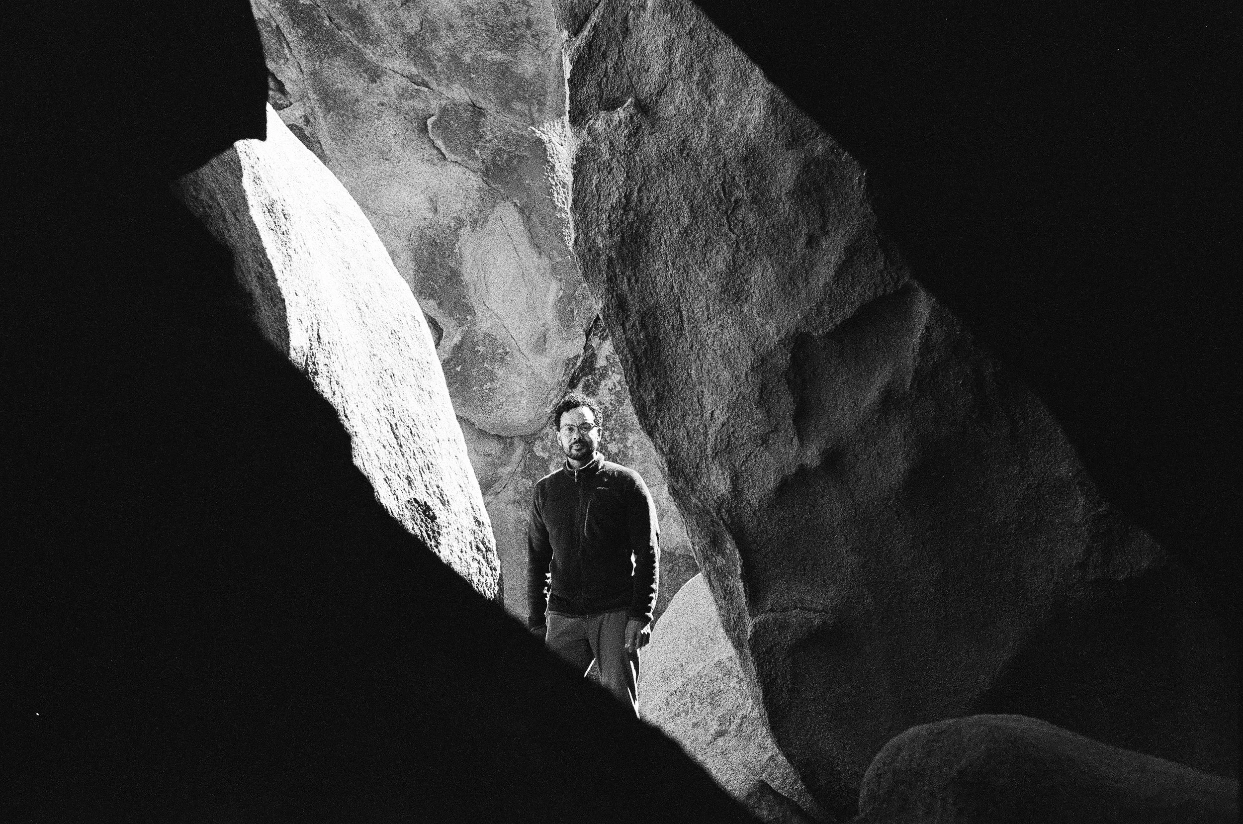 A man standing inside a narrow crevice surrounded by large, textured rocks in a black and white photograph.