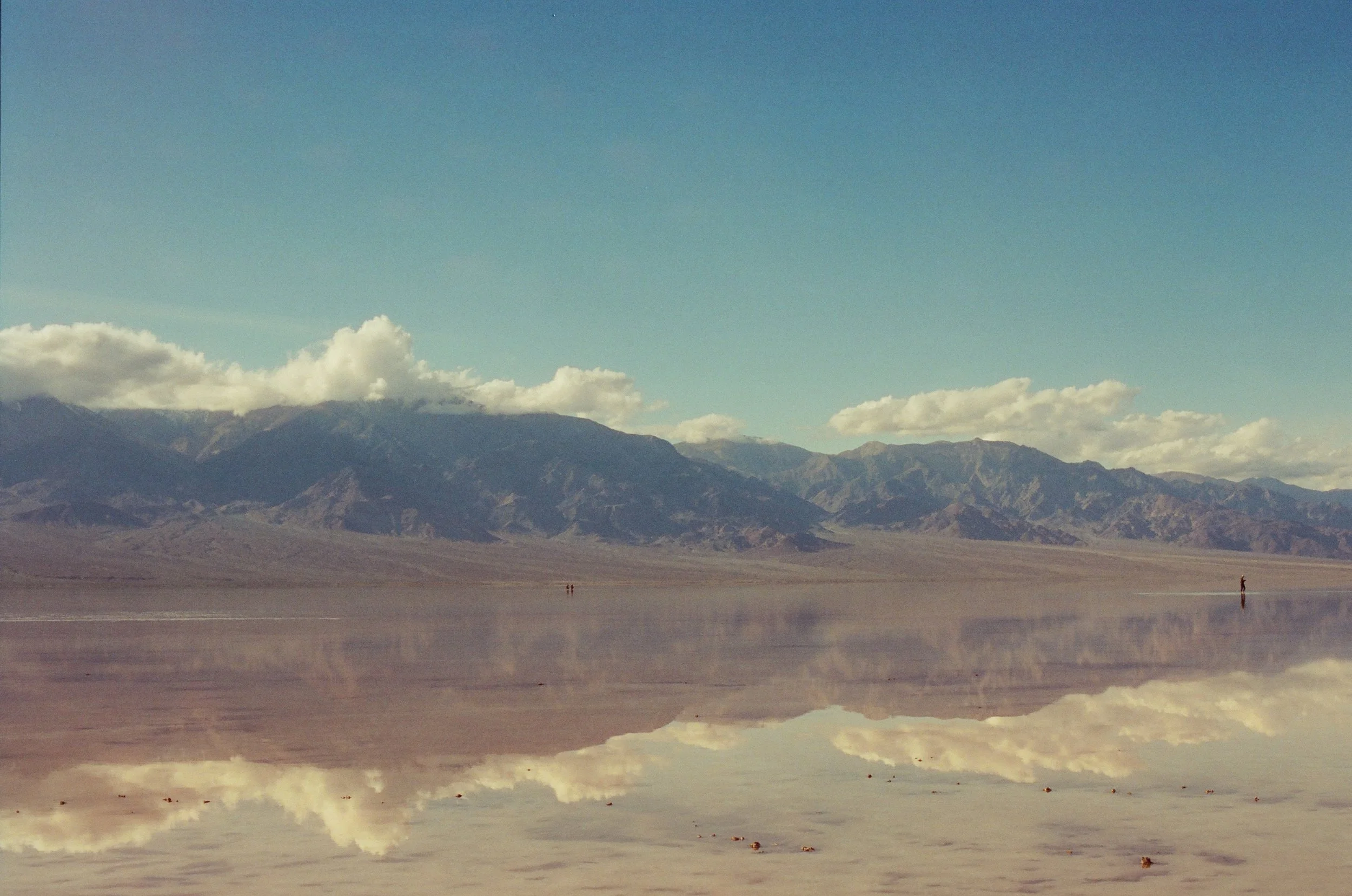 A vast desert landscape with salt flats reflecting the sky and distant mountains, partly cloudy weather.