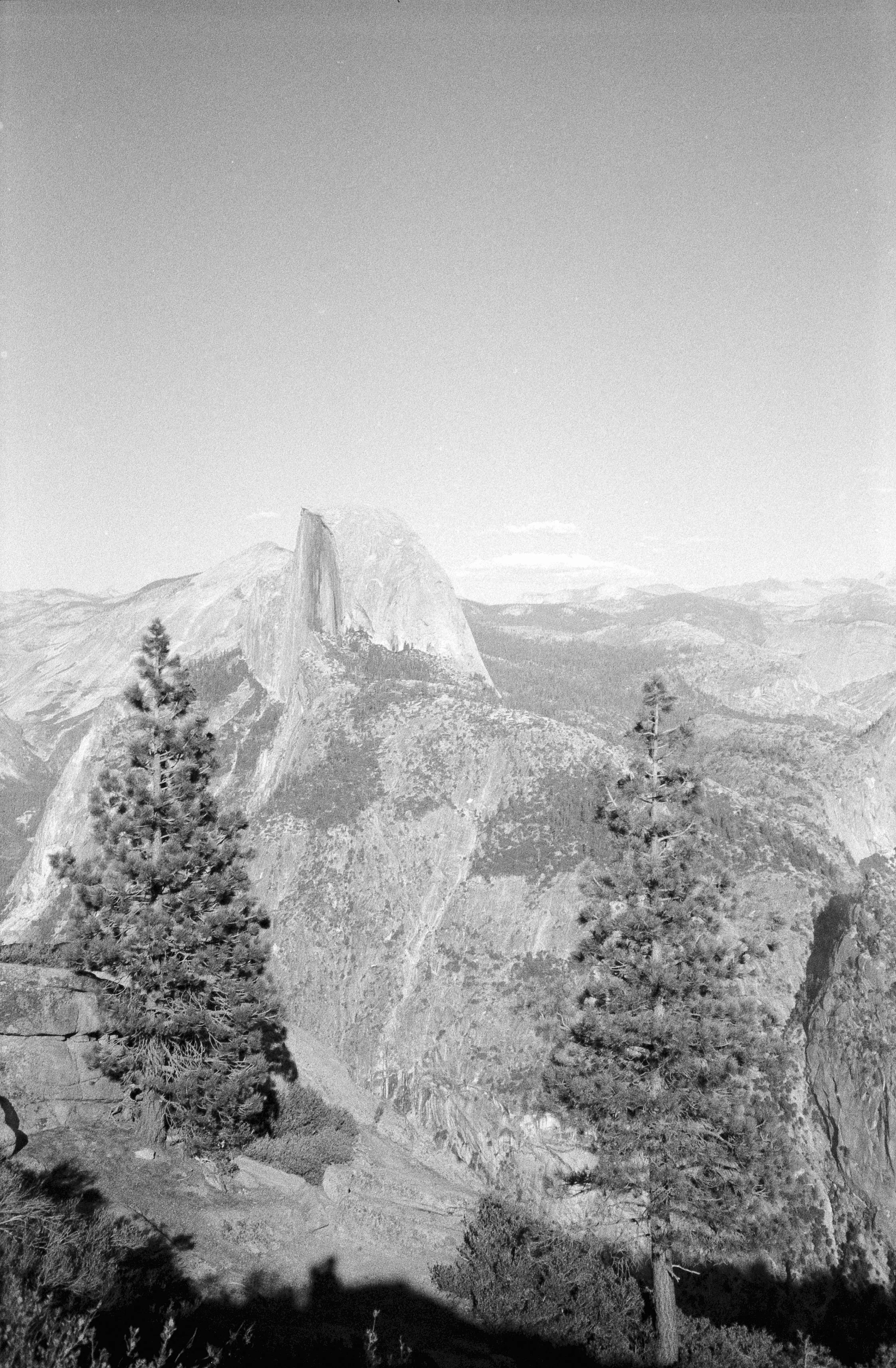 A black and white photograph of a mountainous landscape with a prominent peak, likely Half Dome in Yosemite National Park, surrounded by pine trees and rugged terrain under a clear sky.