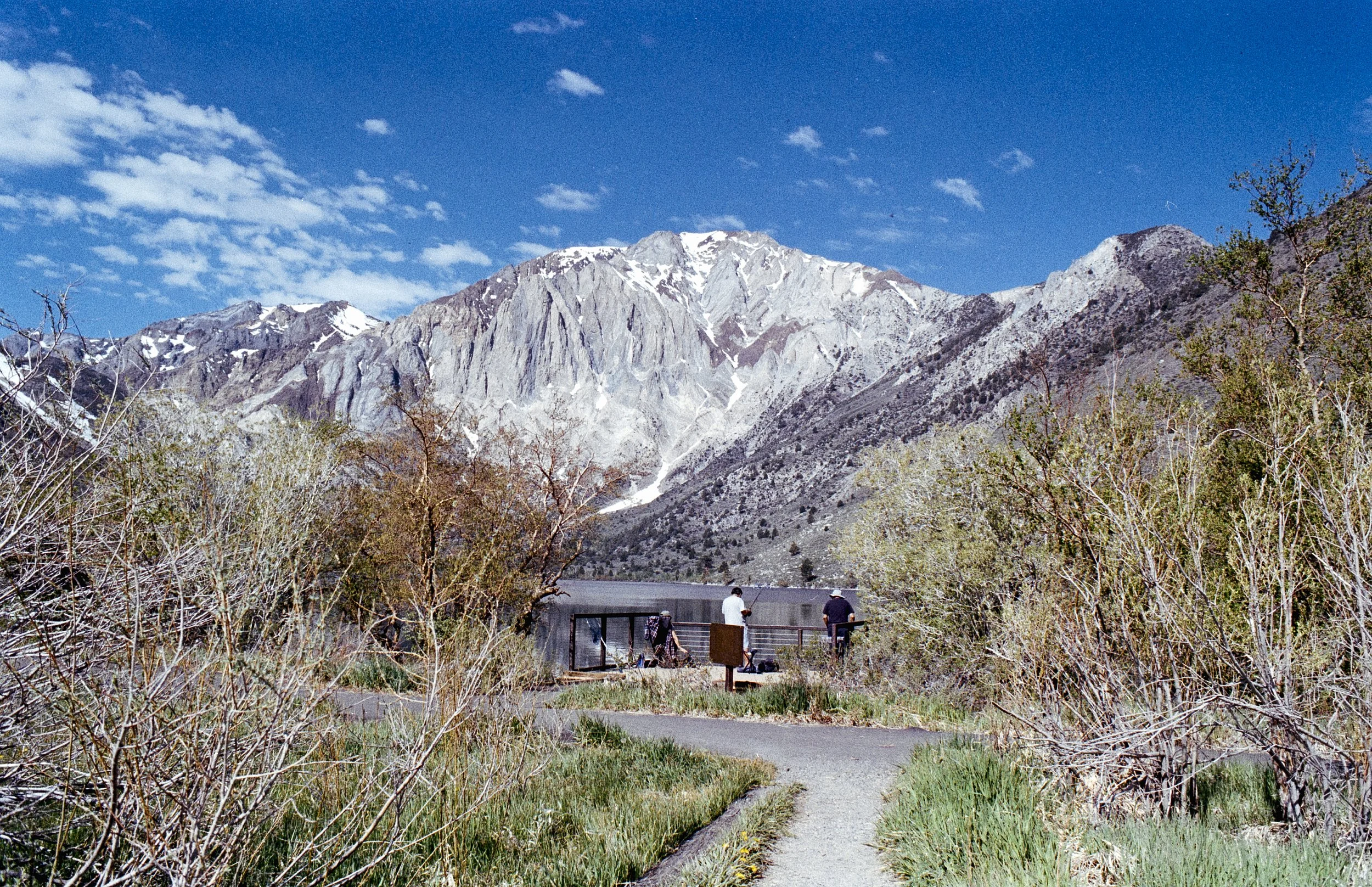 People outdoors near a lake or river with mountains in the background under a partly cloudy blue sky.