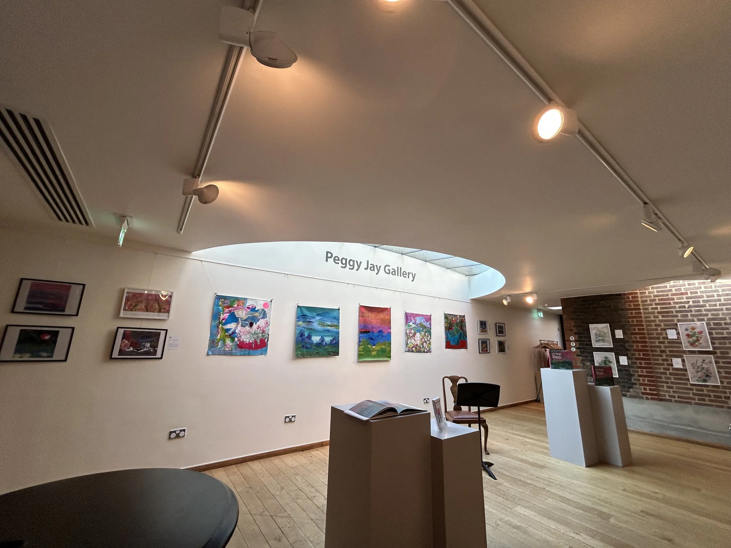 Interior view of an art gallery with colorful paintings displayed on a white wall, a small pedestal with brochures, chairs, and a brick wall in the background. The ceiling has track lighting and a sign indicating 'Peggy Jay Gallery'.