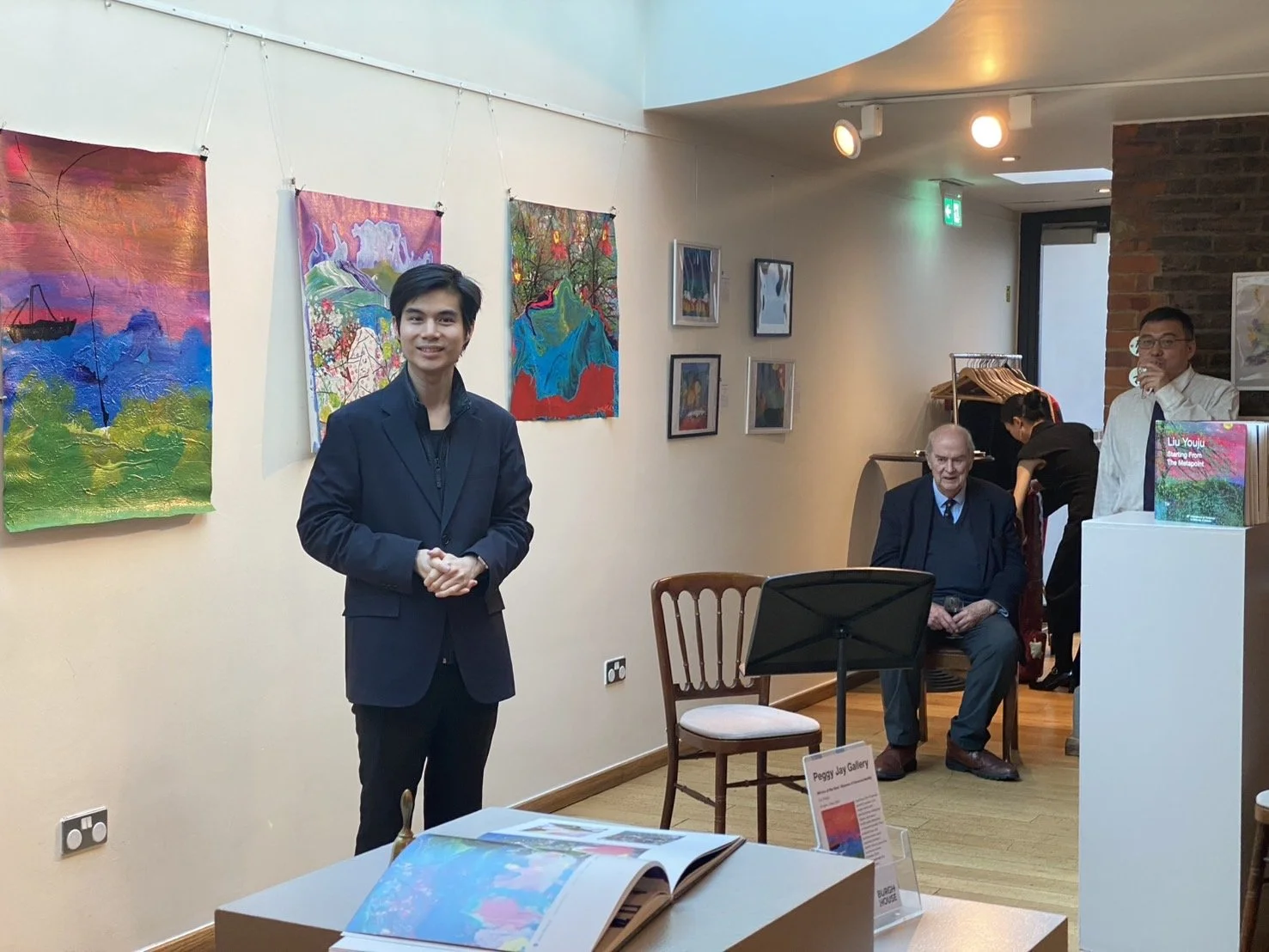 People in an art gallery with colorful paintings on the wall. A man in a black suit is standing and smiling, an older man is sitting on a chair, and a person is working near a clothing rack in the background.