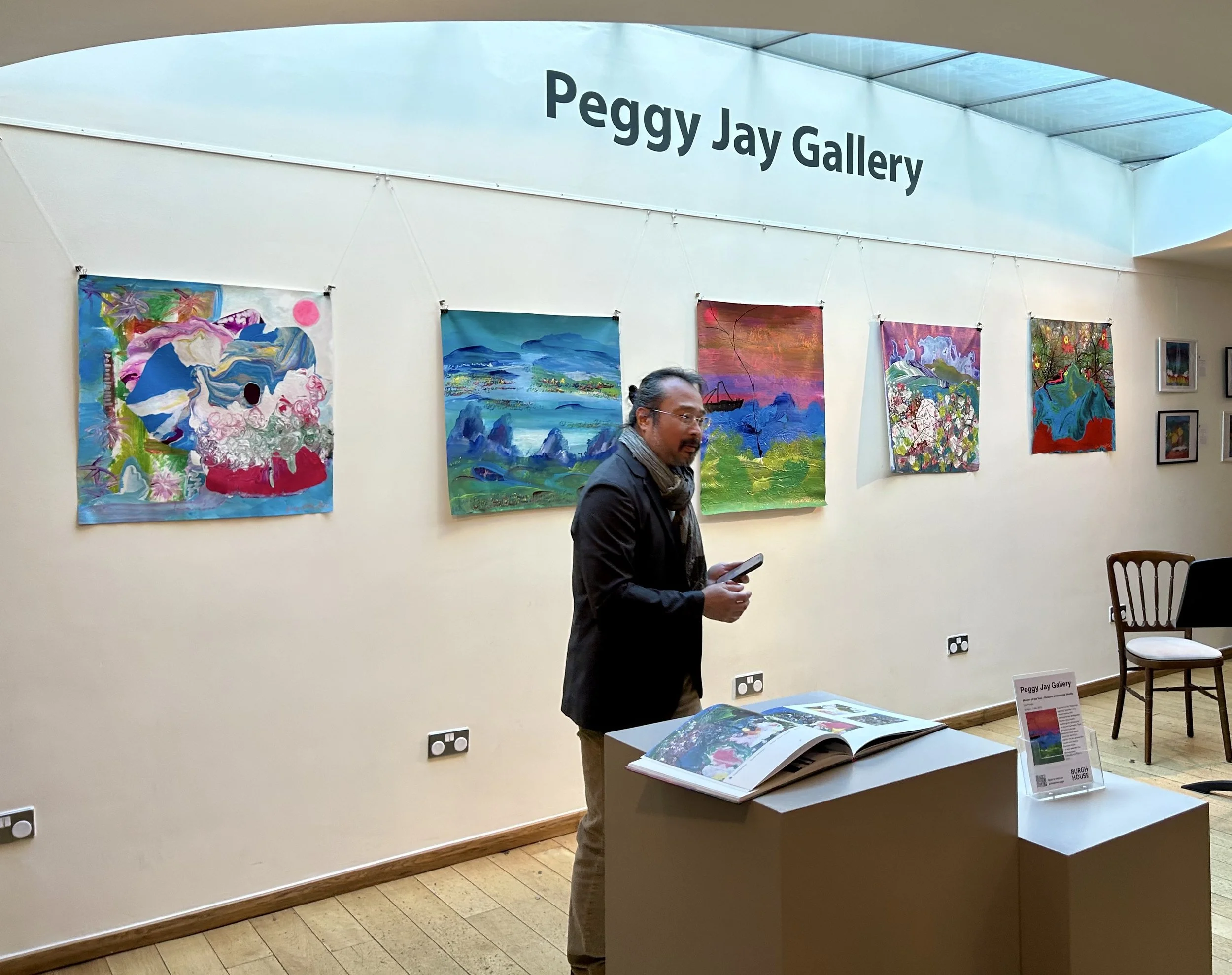 Art exhibit at Peggy Jay Gallery with colorful paintings hanging on the white wall, a man with a scarf holding a phone, informational material on a table, and a sign indicating Peggy Jay Gallery.